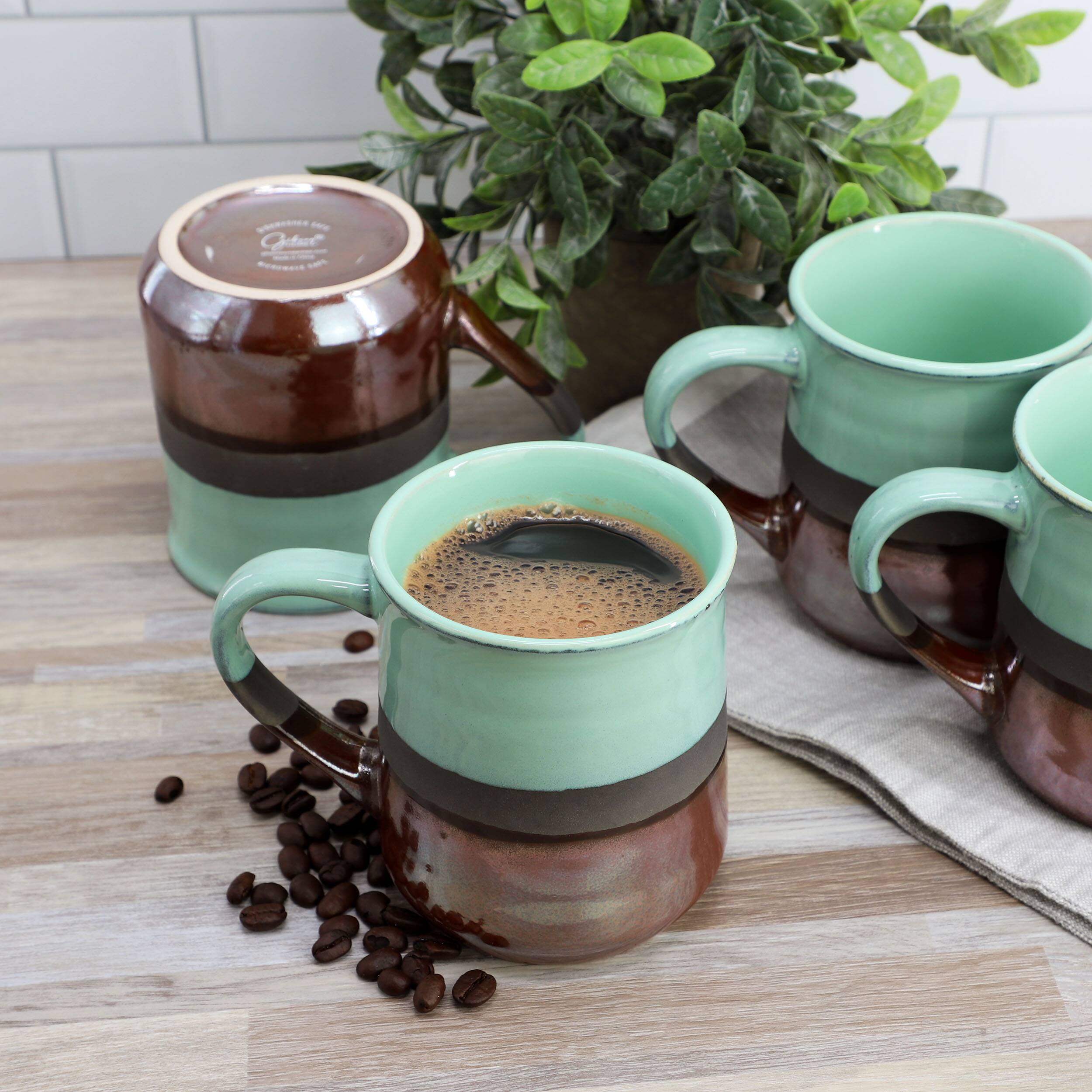 The image features a table with a variety of coffee cups and a coffee pot. There are three coffee cups, one of which is green and black, and the other two are green. The coffee pot is also green and brown. The cups are placed on a table with a potted plant nearby, adding a touch of greenery to the scene. The table appears to be a dining table, and the cups are arranged in a way that suggests they are ready for use.