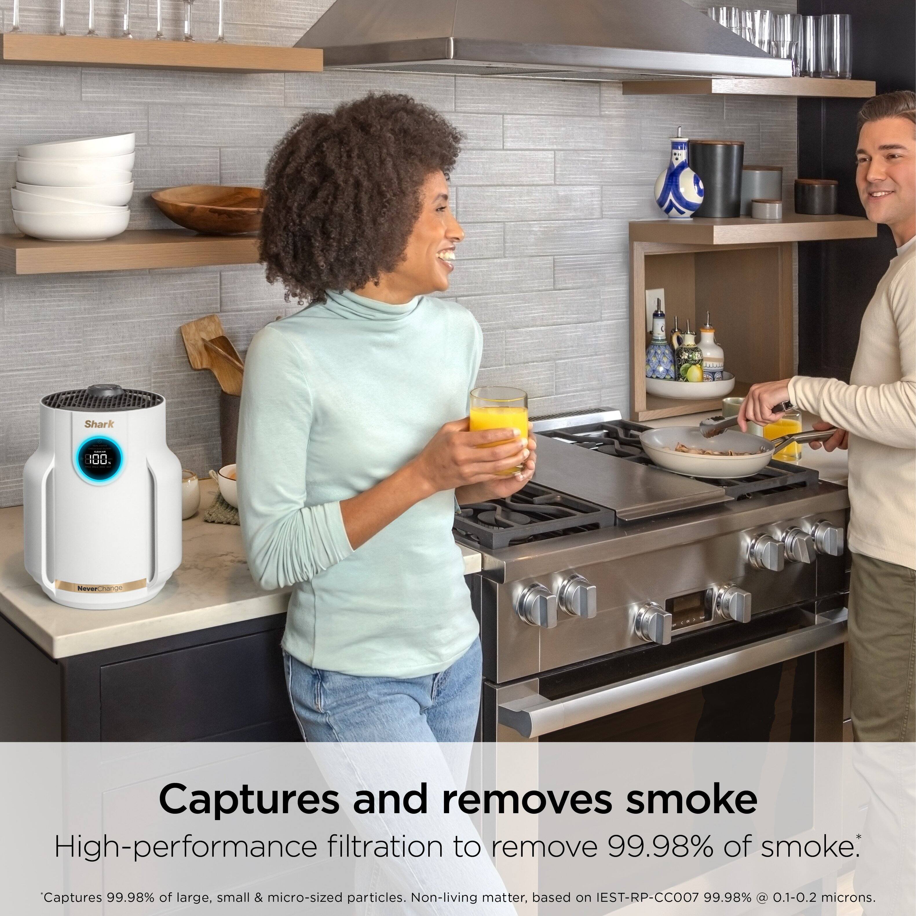 The image features a man and a woman standing in a kitchen. The woman is holding a glass of orange juice, while the man is preparing food in a bowl. The kitchen is well-equipped with various items such as bottles, cups, and a sink. The image also promotes a product that captures and removes smoke, ensuring a high-performance filtration to remove 99.98% of smoke. This product is based on IEST-RP-CC007 and captures 99.98% of large, small, and micro-sized particles, as well as non-living matter.