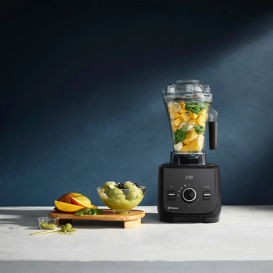 The image features a blender filled with fruits and vegetables, including broccoli, on a countertop. There is also a bowl of fruit and a cup of fruit nearby. The blender is placed on a table, and it is surrounded by various fruits and vegetables. The scene suggests that someone is preparing a healthy meal or smoothie using the blender.
