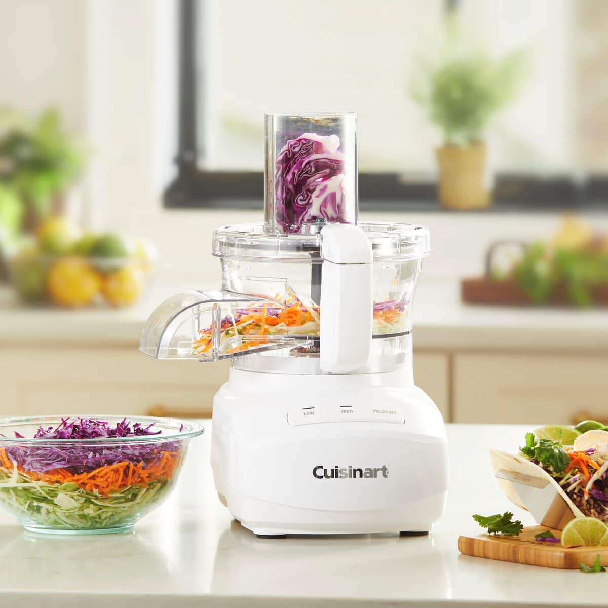 The image shows a Cuisinart food processor on a counter, surrounded by various ingredients such as carrots, onions, and broccoli. The food processor is filled with chopped vegetables, and there are also bowls of food on the counter. The scene suggests that someone is preparing a meal using the Cuisinart food processor.