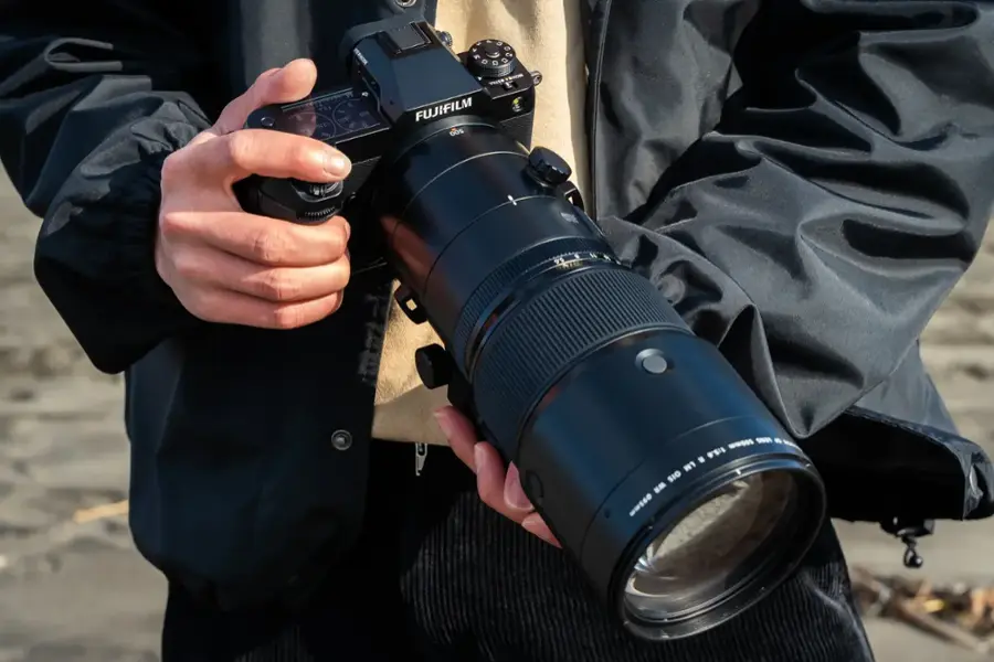 The image features a person holding a camera with a large lens, which is a Fujifilm lens. The lens is quite large and is being held in front of the person's face. The camera is likely being used for photography or videography purposes, given the size of the lens.