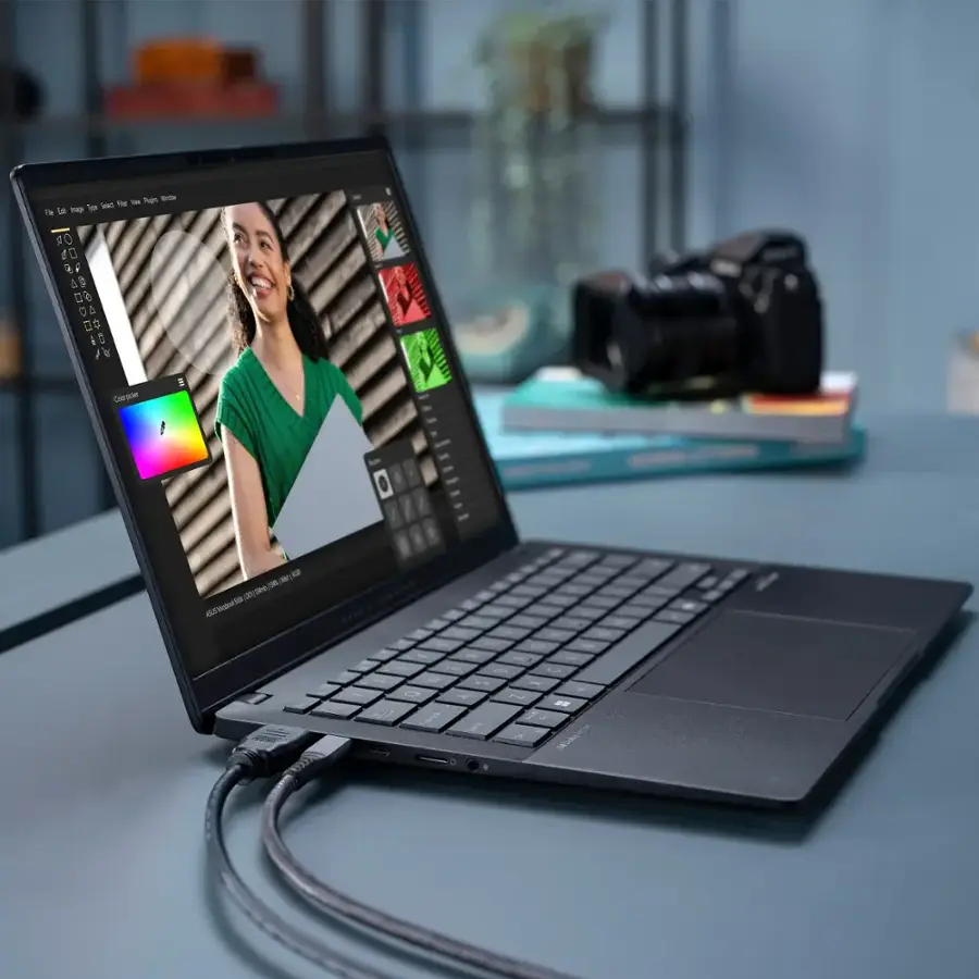 The image shows a laptop computer with a picture of a woman on the screen. The laptop is sitting on a desk, and there are several books nearby. Additionally, there is a camera placed next to the laptop, and a potted plant is located in the background. The laptop is open, and it appears to be in use.