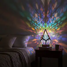 The image shows a colorful, kaleidoscopic light projection on a wall, emanating from a decorative lamp placed on a bedside table. The lamp has a geometric, faceted design and is casting vibrant, multicolored patterns across the room. On the table, there is a white mug and a stack of books. The scene is set in a cozy bedroom with a bed covered in a textured blanket and pillows.