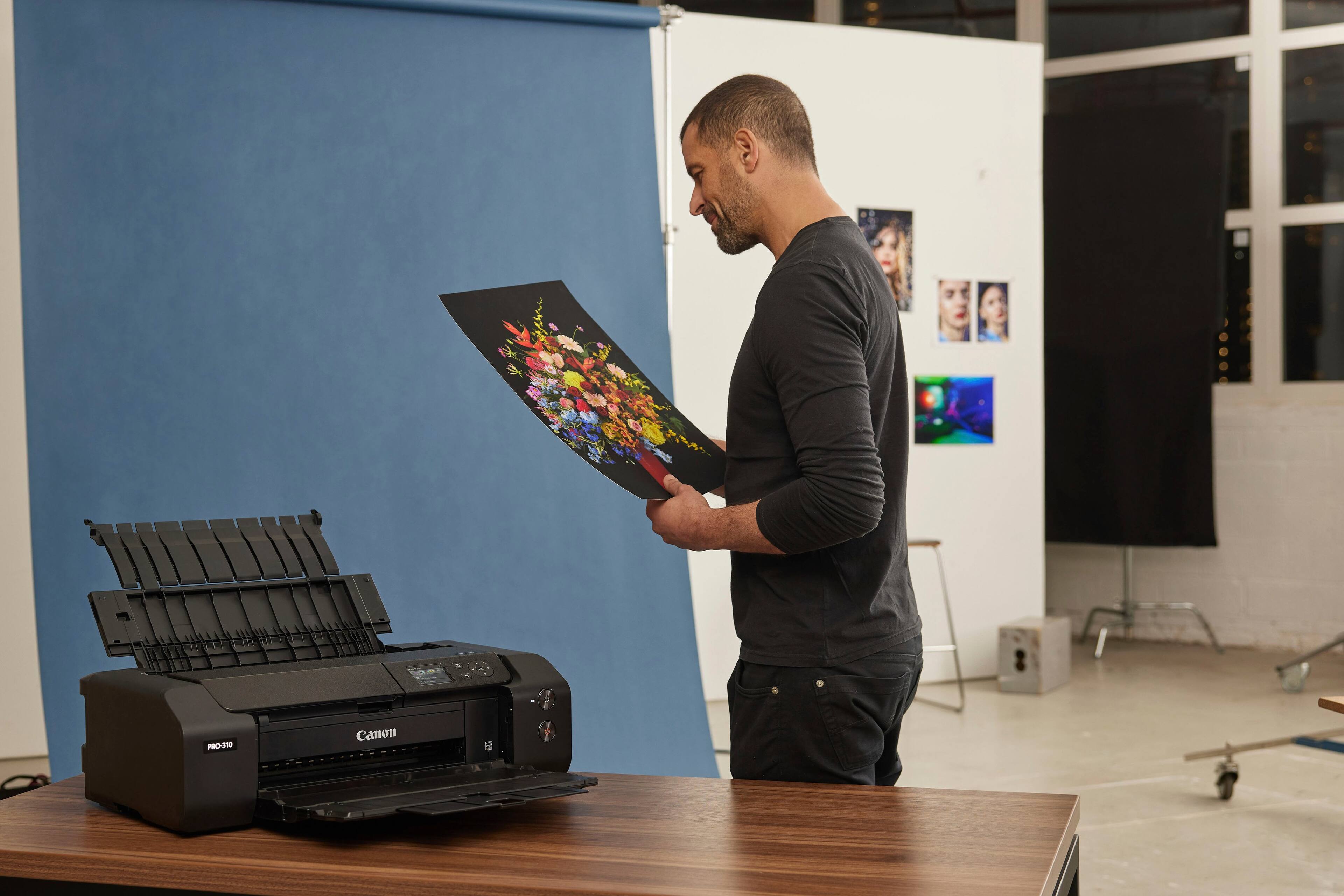 The man is holding a colorful printout in front of a Canon printer.