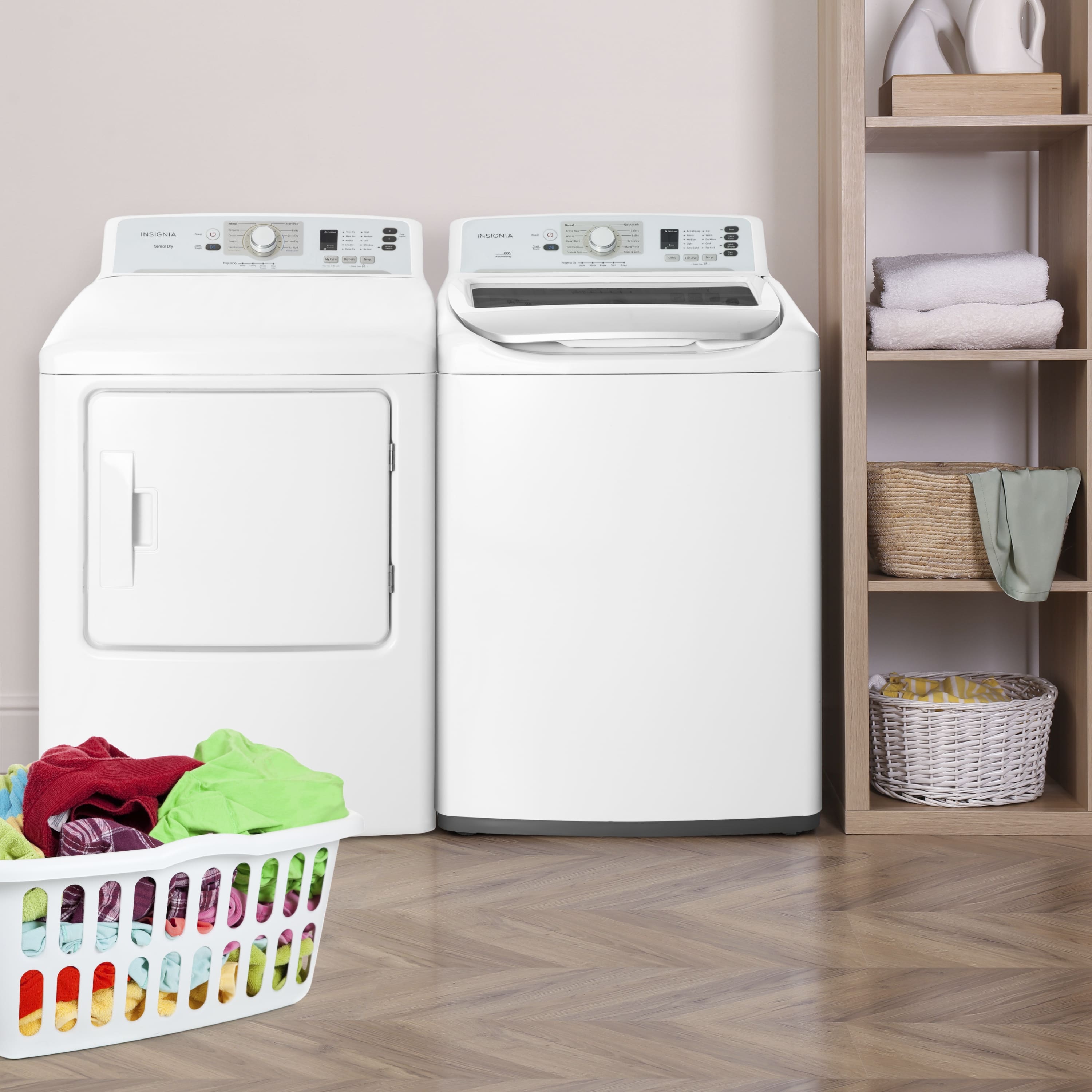 The image shows a laundry room with two white washing machines. One of the machines has a basket of clothes next to it, while the other machine is empty. The room also contains a dryer and a shelf with towels. The scene is set in a home, and the washing machines are placed on the floor.