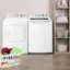 The image shows a laundry room with two white washing machines. One of the machines has a basket of clothes next to it, while the other machine is empty. The room also contains a dryer and a shelf with towels. The scene is set in a home, and the washing machines are placed on the floor.