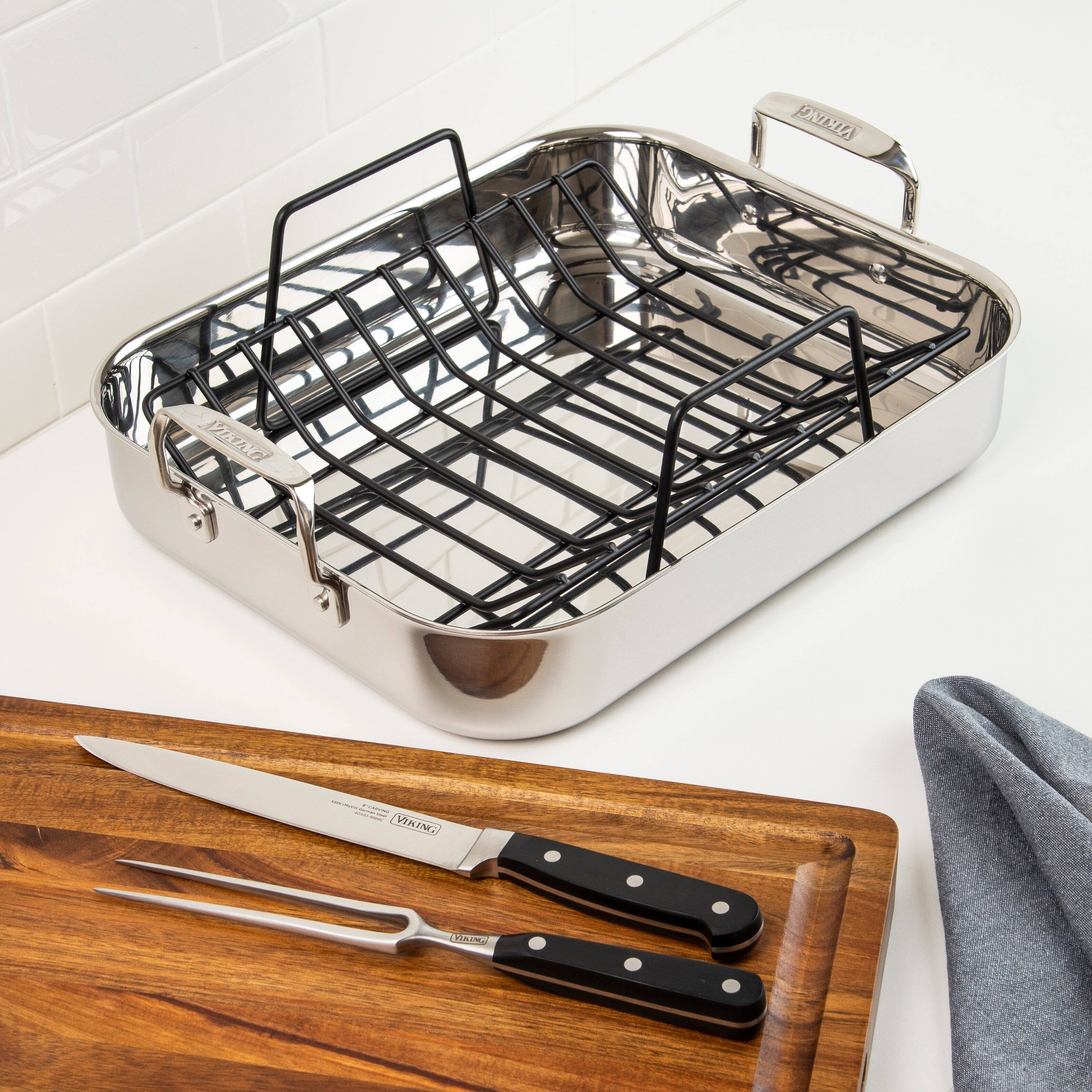 The image shows a stainless steel grill pan with a black grill on a white countertop. Next to the pan, there are two knives, one of which is a large knife, and a pair of tongs. The knives and tongs are placed on a wooden cutting board.
