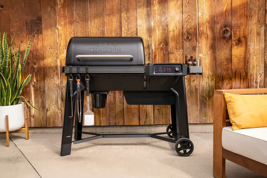 Traeger Grill is a type of grill that is designed for outdoor cooking. It is a black grill with a stainless steel finish and is equipped with a digital control panel. The grill is placed on a wooden deck, and there is a potted plant nearby.