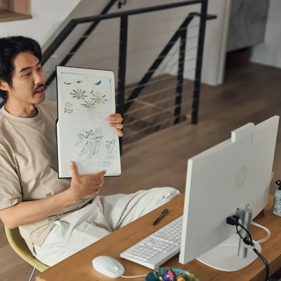 The man is sitting at a desk with a laptop and a book open in front of him. He is holding a book with a drawing of a banana in it. The laptop is on the right side of the desk, and the man is sitting on a chair.