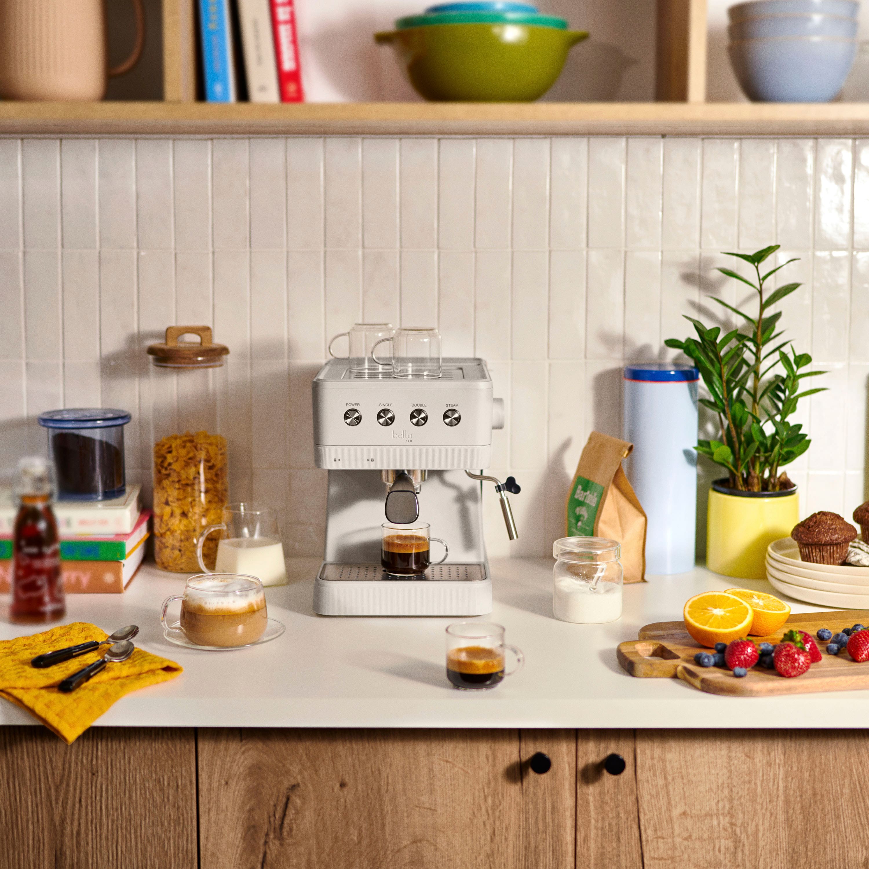 The image features a kitchen counter with various food items and kitchen appliances. There is a coffee maker, a blender, and a juicer on the counter. There are also several cups, bowls, and bottles placed around the counter. Additionally, there is a potted plant and a book on the counter. The counter is filled with food, including a bowl of fruit and a plate of pastries.