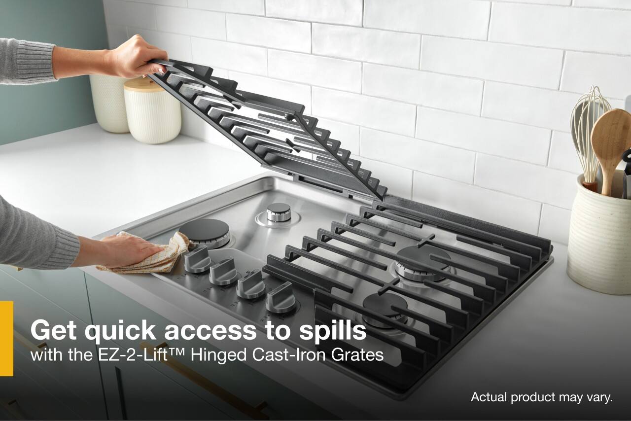 The image features a person opening a kitchen sink, which is filled with various kitchen utensils such as knives, spoons, and a bowl. The sink is surrounded by a white tile wall. The advertisement suggests that the EZ-2-LiftTM TM Cast-Iron Grates can provide quick access to spills, making it easier to clean up and maintain a hygienic kitchen environment.