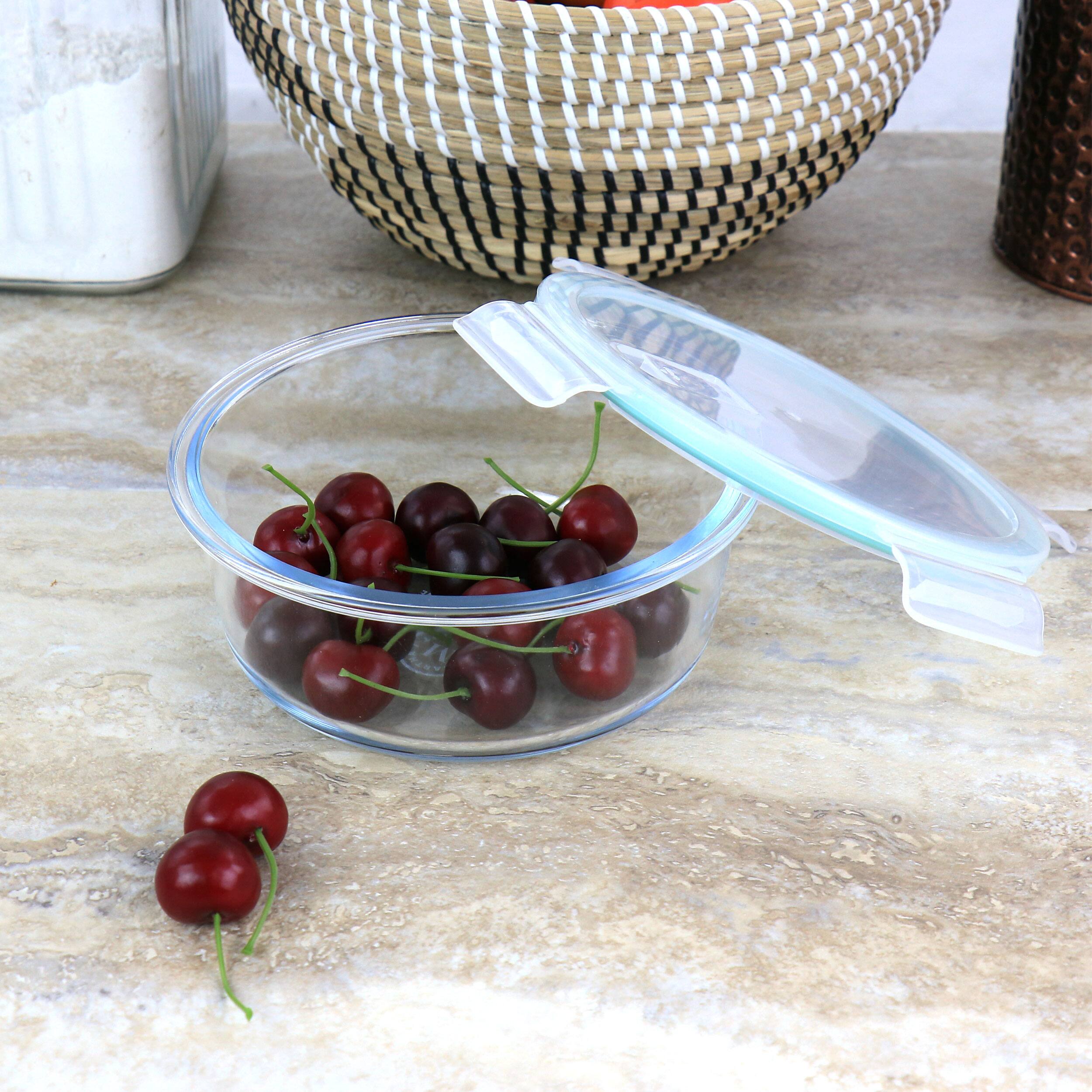 The image features a clear plastic container filled with cherries, with the lid open. The container is placed on a countertop, and there are additional cherries scattered around the container. The scene also includes a bowl and a bottle, which are placed nearby.
