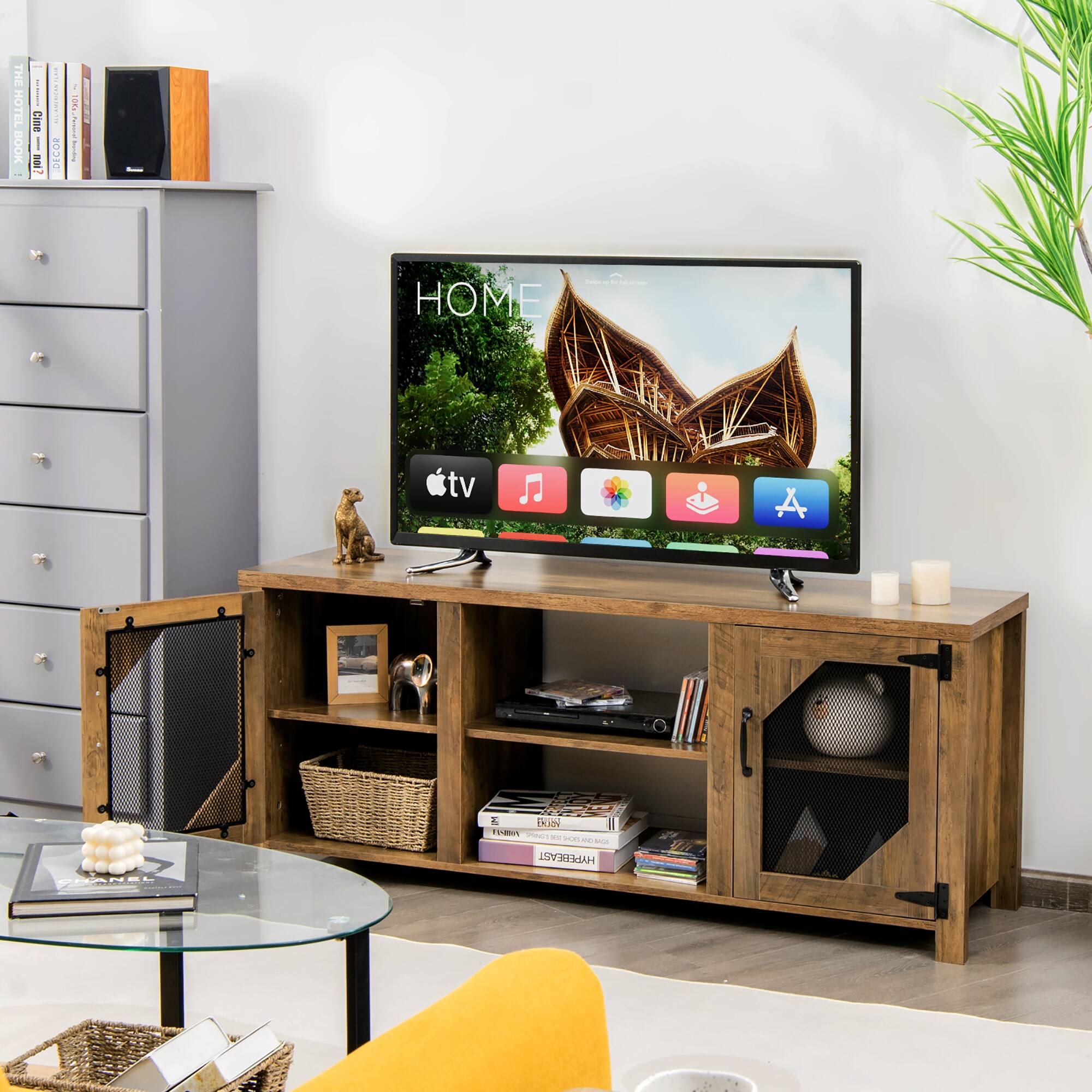 The image shows a living room with a flat screen TV on a wooden stand. The TV is displaying the Apple TV menu, which includes options for music, movies, and TV shows. There is a remote control placed nearby, and a bookshelf with various books is located in the room. Additionally, there is a potted plant and a vase, adding to the cozy atmosphere of the living space.