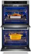 The image shows a blue oven with two dishes inside. One dish is a yellow bowl, and the other is a silver pan. The oven is open, revealing the food being cooked. The time displayed on the oven is 12:03.