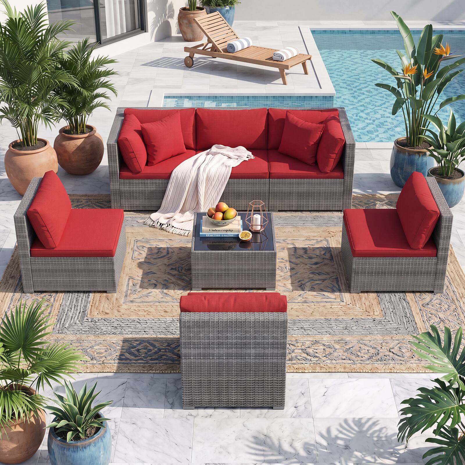 A cozy outdoor patio area features a wicker sofa set with vibrant red cushions, a matching ottoman, and a wooden sun lounger. The space is adorned with potted plants and a small table holding a book titled "A Little Crafty" and a bowl of fruit.