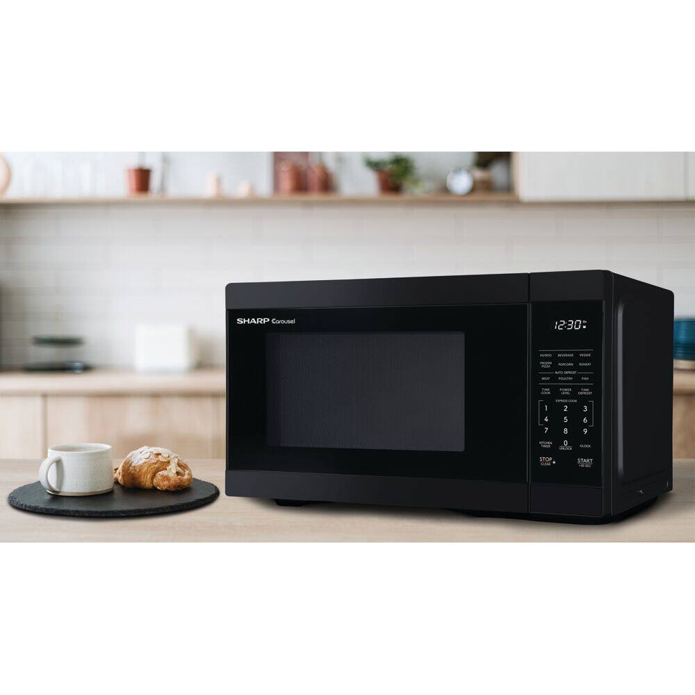 The image features a black Sharp microwave oven sitting on a wooden counter. Next to the microwave, there is a plate with a pastry on it. The microwave is placed on a kitchen counter, surrounded by various items such as a cup, a bowl, and a potted plant. The scene suggests a cozy and functional kitchen space.