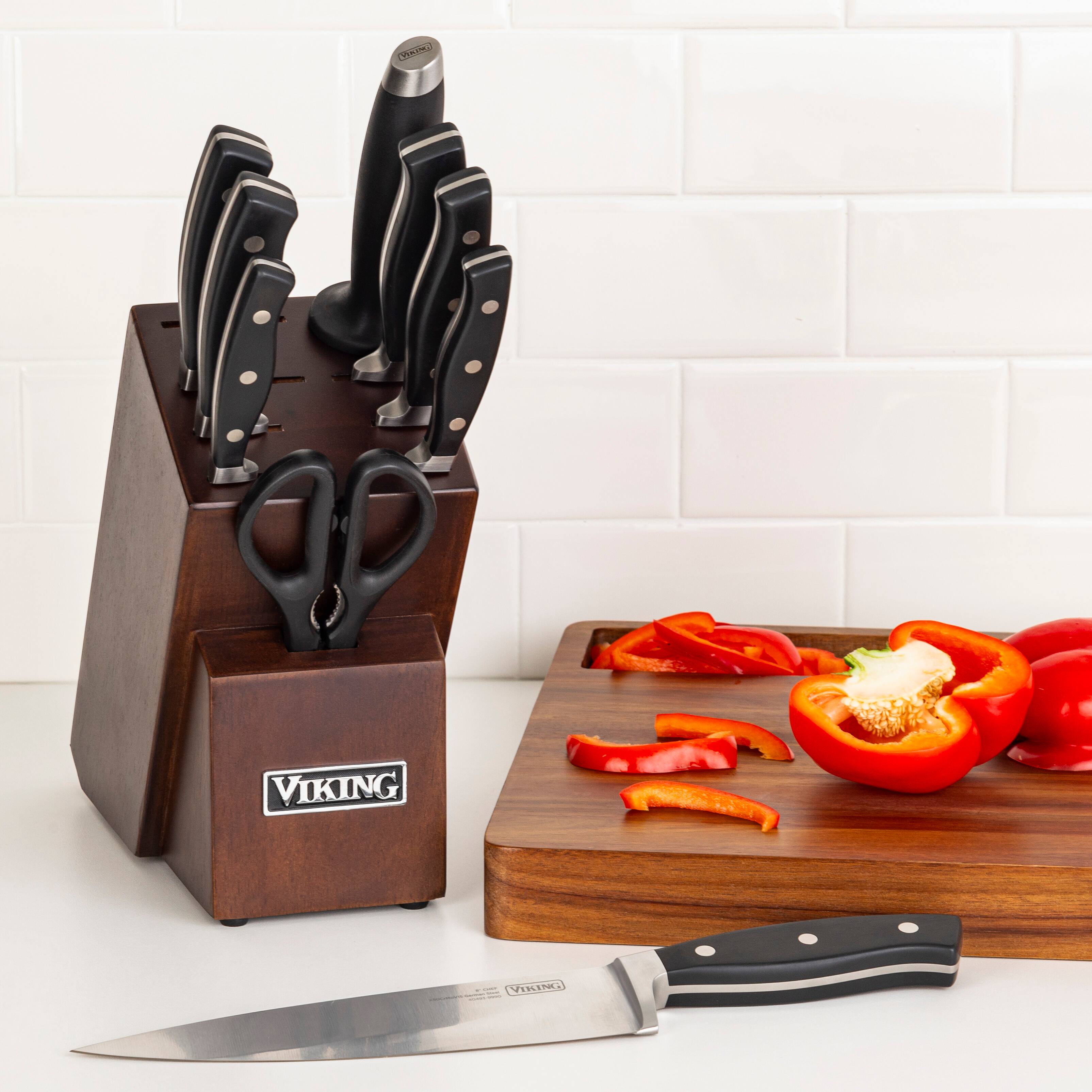 The image features a wooden cutting board with a knife and a pair of scissors on it. The knife is placed on the left side of the cutting board, while the scissors are located on the right side. The cutting board is placed on a white countertop, and there are several peppers nearby, possibly indicating that they are being prepared for a meal. The presence of the knife and scissors suggests that this is a kitchen setting, and the cutting board is being used for food preparation.