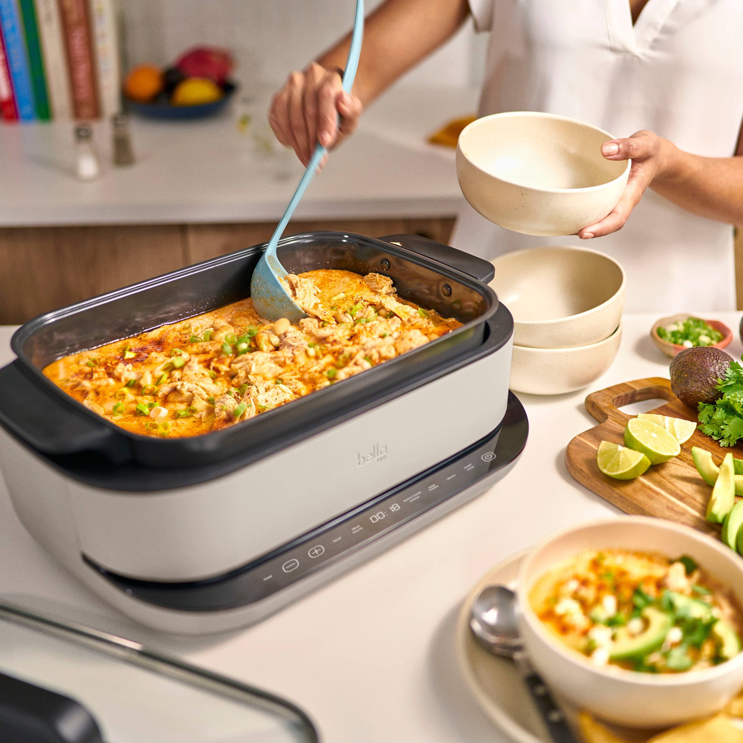 The image shows a woman standing in a kitchen, holding a bowl and a spoon. She is serving food from a crockpot, which is placed on a counter. The counter also has a variety of bowls and a cup. There are several books scattered around the kitchen, possibly cookbooks or other reading materials. Additionally, there are two oranges on the counter, adding a touch of color to the scene.