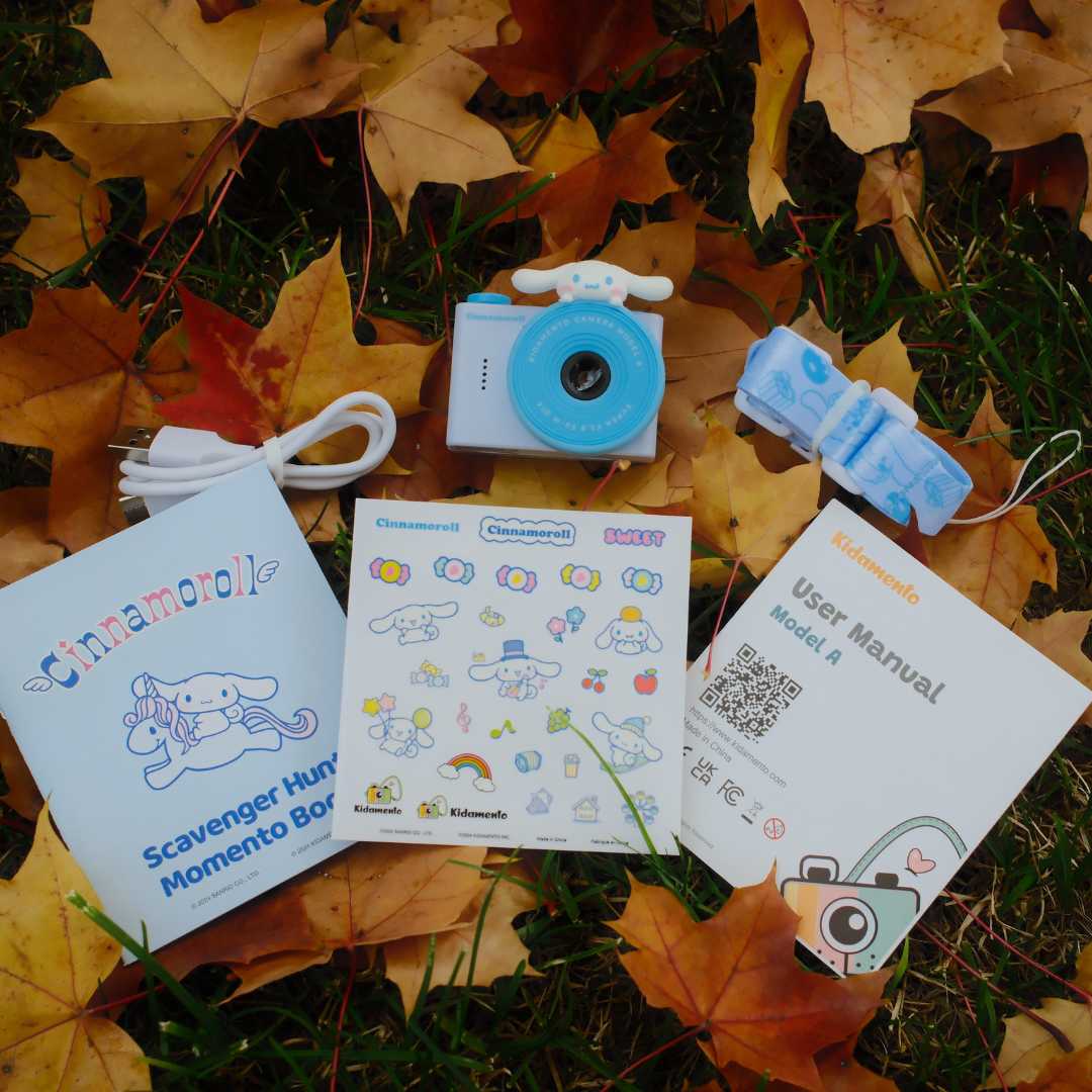 The image features a variety of items, including a camera, a book, and a manual. The camera is placed on top of the book, which is titled "Cinnamoroll." The manual is also present, providing instructions for the camera. The items are arranged on a grassy surface, with leaves scattered around them.