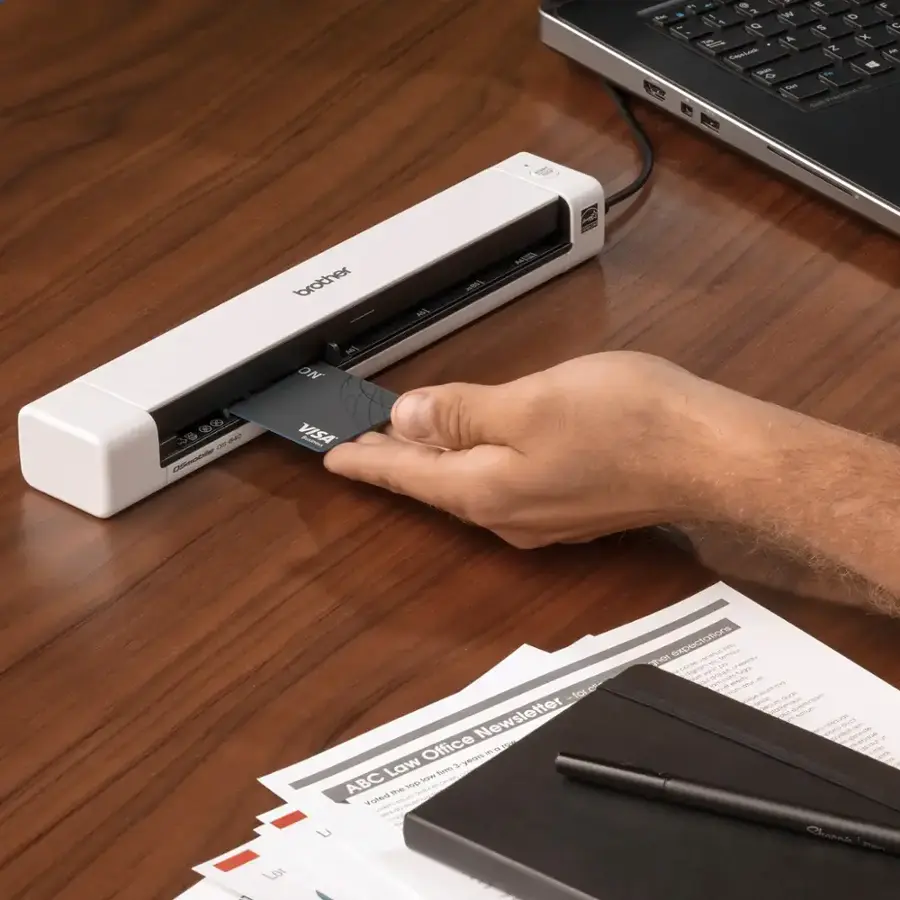 The image shows a person using a printer to print out a Visa card. The printer is placed on a wooden desk, and the person is holding the card in their hand. The scene also includes a laptop and a keyboard on the desk. The text on the image is not coherent and does not provide any additional context or information about the scene.