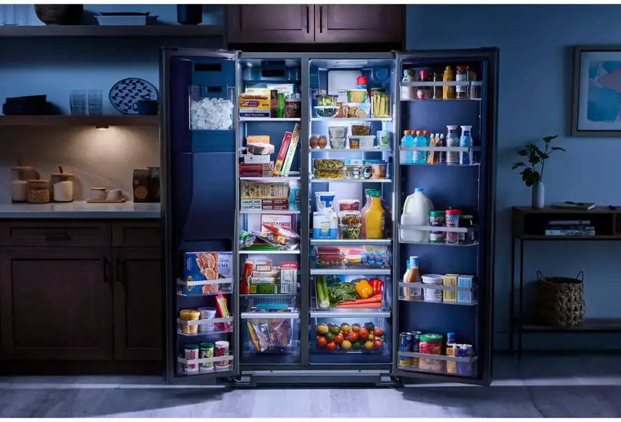 The image shows a refrigerator with its light on, revealing a well-stocked interior. The refrigerator is filled with various food items, including several bottles, fruits, and vegetables. There are also numerous books placed inside the refrigerator, which is an unusual sight. The refrigerator is located in a kitchen with a dining table nearby. The scene is set at night, giving the refrigerator a spotlight and emphasizing its contents.
