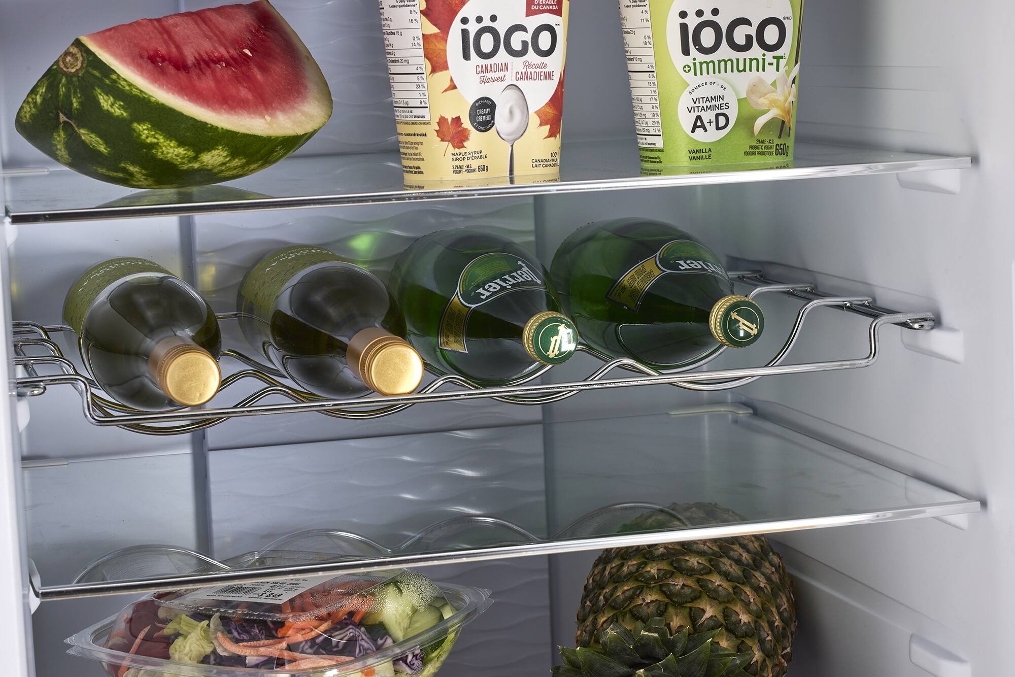 The image shows a refrigerator with various food items and drinks. There are two bottles of wine, a bottle of juice, and a bottle of water. Additionally, there is a container of fruit, a container of vegetables, and a container of yogurt. The refrigerator also has a shelf with a watermelon and a pineapple. The refrigerator is stocked with a variety of food and drinks, providing a diverse selection of items for consumption.