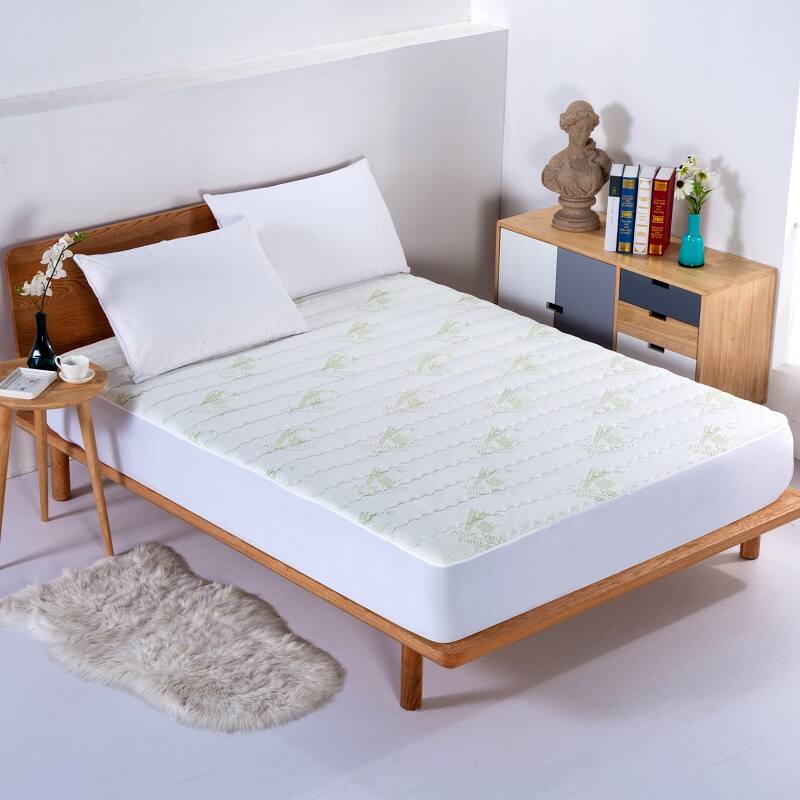 The image shows a neatly arranged bedroom with a wooden bed frame and a mattress covered with a white sheet featuring a subtle leaf pattern. There are two white pillows on the bed. On the left side, there is a small wooden bedside table with a black vase holding white flowers and a book. On the right side, there is a wooden nightstand with a statue, a stack of books, and a turquoise vase with white flowers. A fluffy gray rug is placed on the floor near the bed.