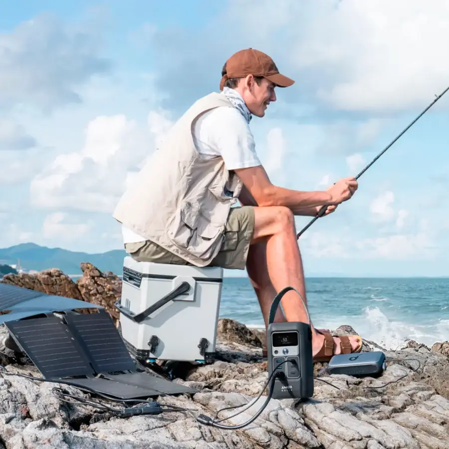 "A man is sitting on a cooler and fishing on a rocky shore. He is wearing a brown hat and a white shirt. The man is holding a fishing rod and appears to be enjoying his time outdoors."