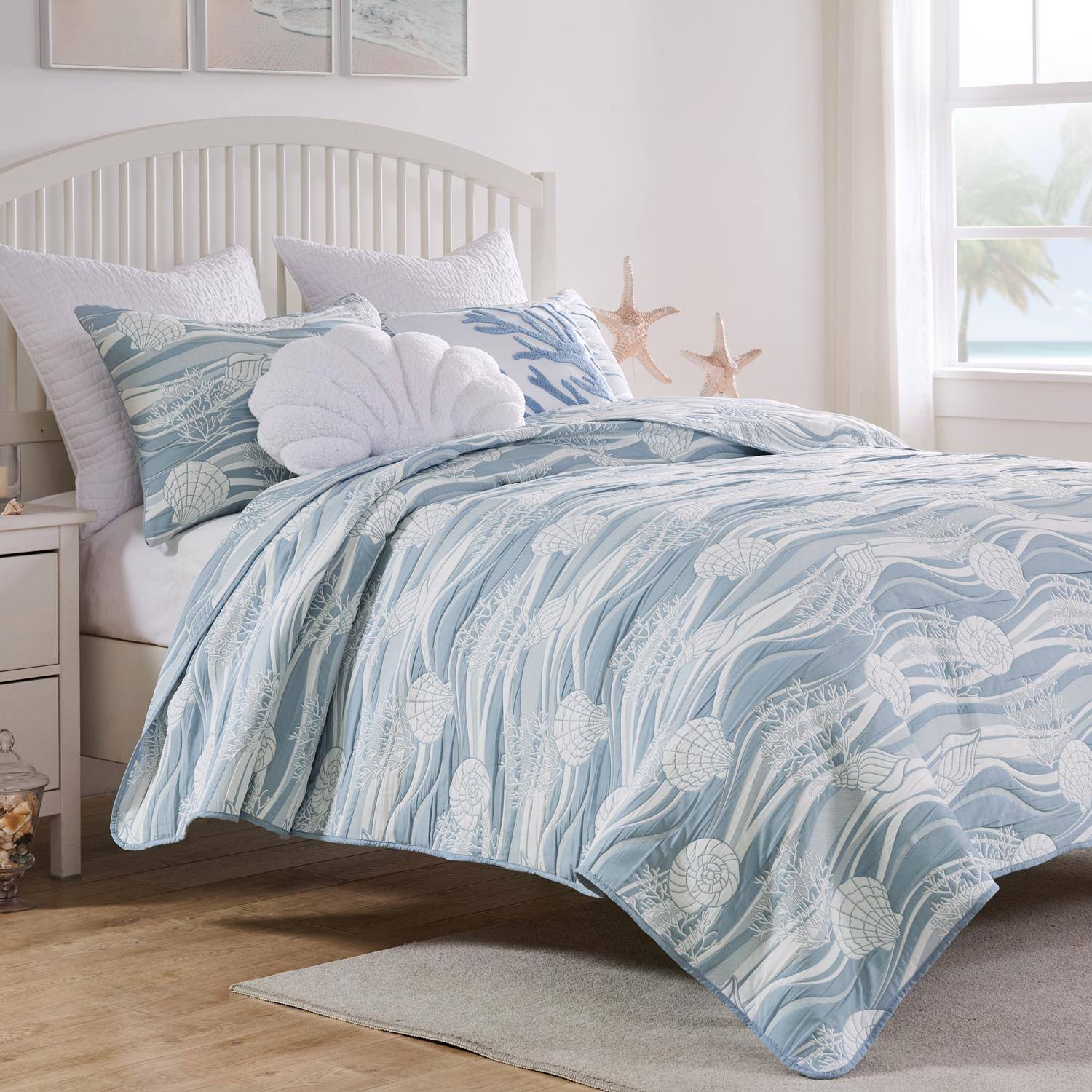 The image shows a neatly made bed with a light blue and white patterned comforter featuring seashell designs. The bed has multiple pillows, including a decorative one with a large white shell. The room has a coastal theme, with starfish decorations on the wall and a window showing a view of palm trees and the ocean. The furniture is light-colored, and the floor is wooden.