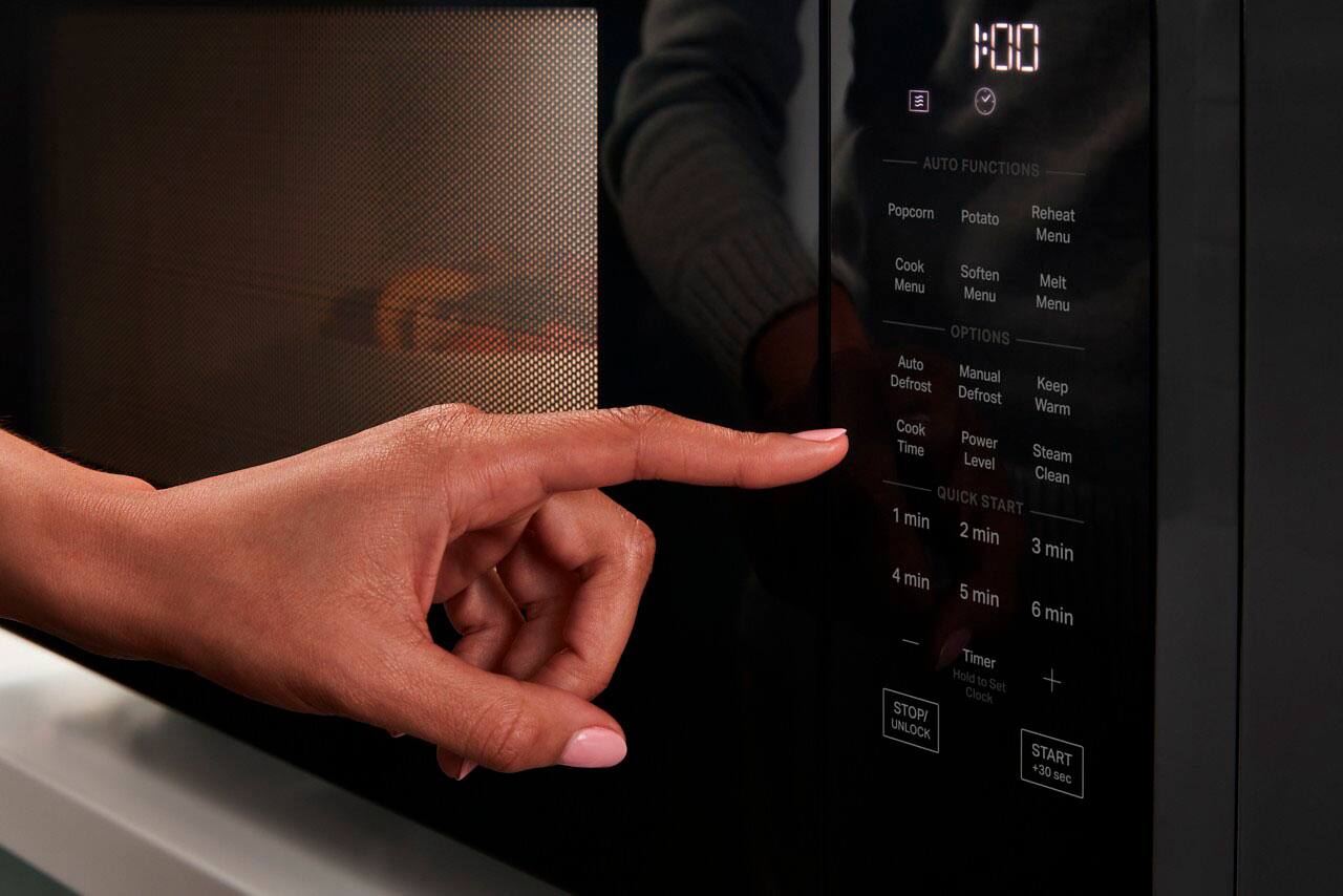The image shows a person using a microwave oven, with the time displayed as 1:00. The microwave has several buttons, including options for auto functions, such as popcorn, potato reheat, and melt. Other menu options include cook, soften, and keep warm. The user can also adjust the cook time, power level, and steam clean settings. Additionally, the microwave has a timer function and a quick start option. The person's hand is pointing at the buttons, indicating their selection of the desired function.
