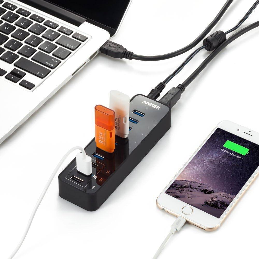 In a photo, a laptop is connected to a black Anker power hub with multiple ports. A smartphone shows "100% Charged" on its screen.