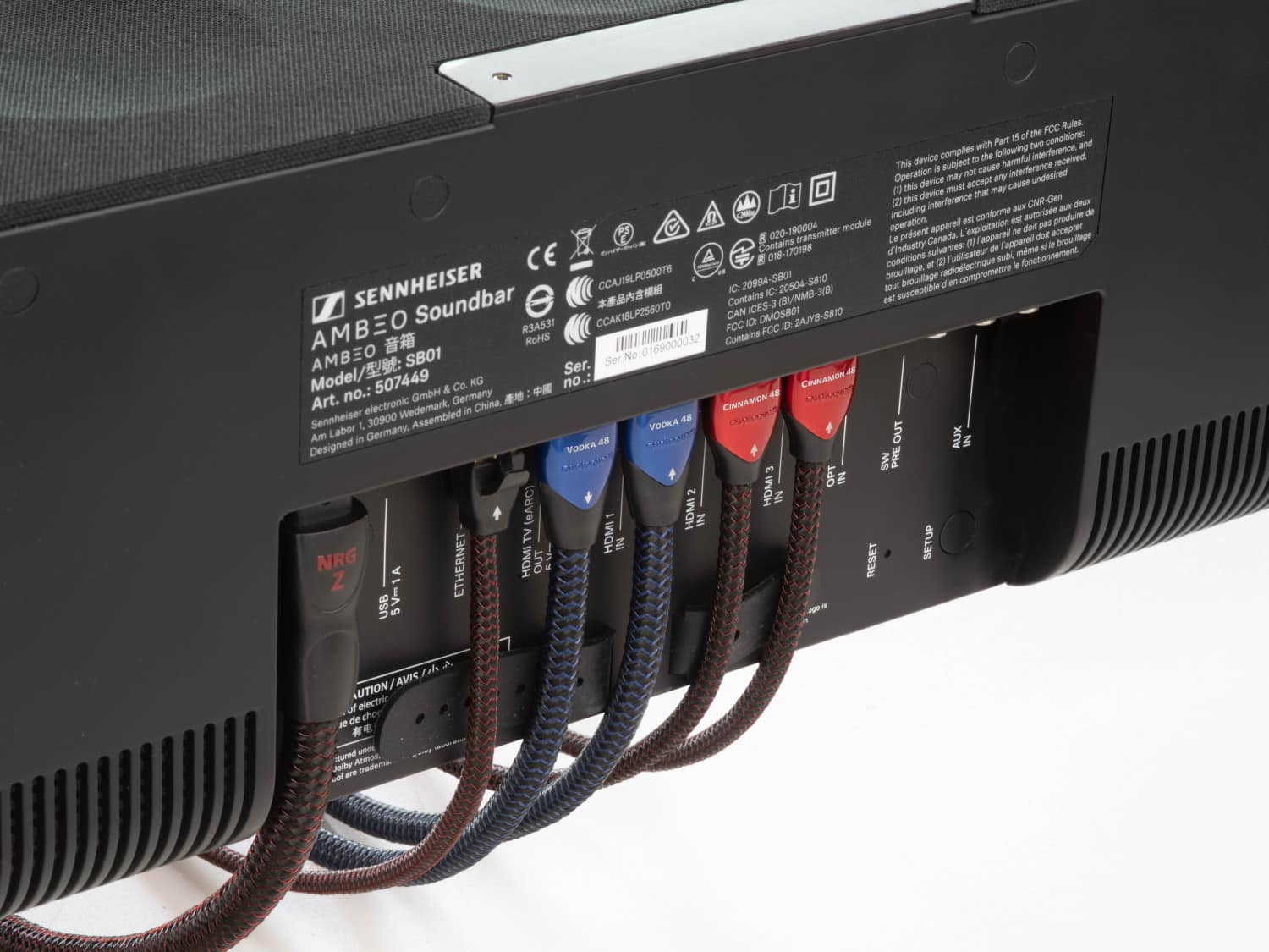 The image shows a Sennheiser soundbar with a back panel that has a sticker on it. The sticker contains information about the device, such as its model number, serial number, and FCC rules. The soundbar is connected to a computer, and there are several cords plugged into the back of the device. The cords are likely used for power, audio input, or other functions. The soundbar is designed to enhance the audio experience for users, providing a more immersive and enjoyable listening experience.