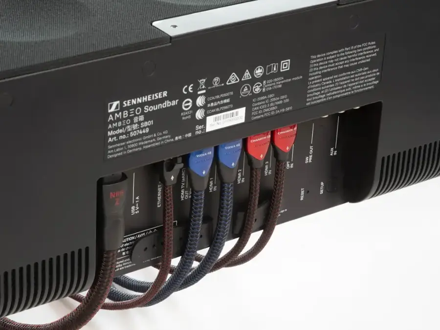 The image shows a Sennheiser soundbar with a back panel that has a sticker on it. The sticker contains information about the device, such as its model number, serial number, and FCC rules. The soundbar is connected to a computer, and there are several cords plugged into the back of the device. The cords are likely used for power, audio input, or other functions. The soundbar is designed to enhance the audio experience for users, providing a more immersive and enjoyable listening experience.