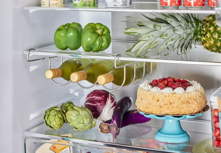 The refrigerator is filled with a variety of food items, including a cake, vegetables, and bottles of wine. The cake is placed on a blue plate, and the vegetables are stored in a crisper drawer. There are also several bottles of wine, with one placed on the top shelf and another on the middle shelf. The refrigerator is well-stocked and ready for use.