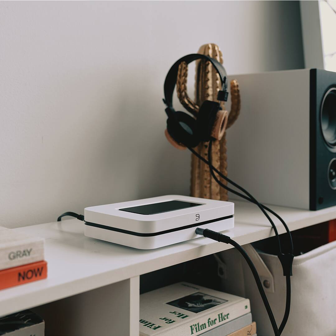 The image features a white shelf with a variety of items on it. There is a small white device, possibly a speaker or a router, sitting on the shelf. Next to the device, there is a pair of headphones. Additionally, there are several books on the shelf, with some of them placed in a stack. A cactus is also present on the shelf, adding a touch of greenery to the scene.