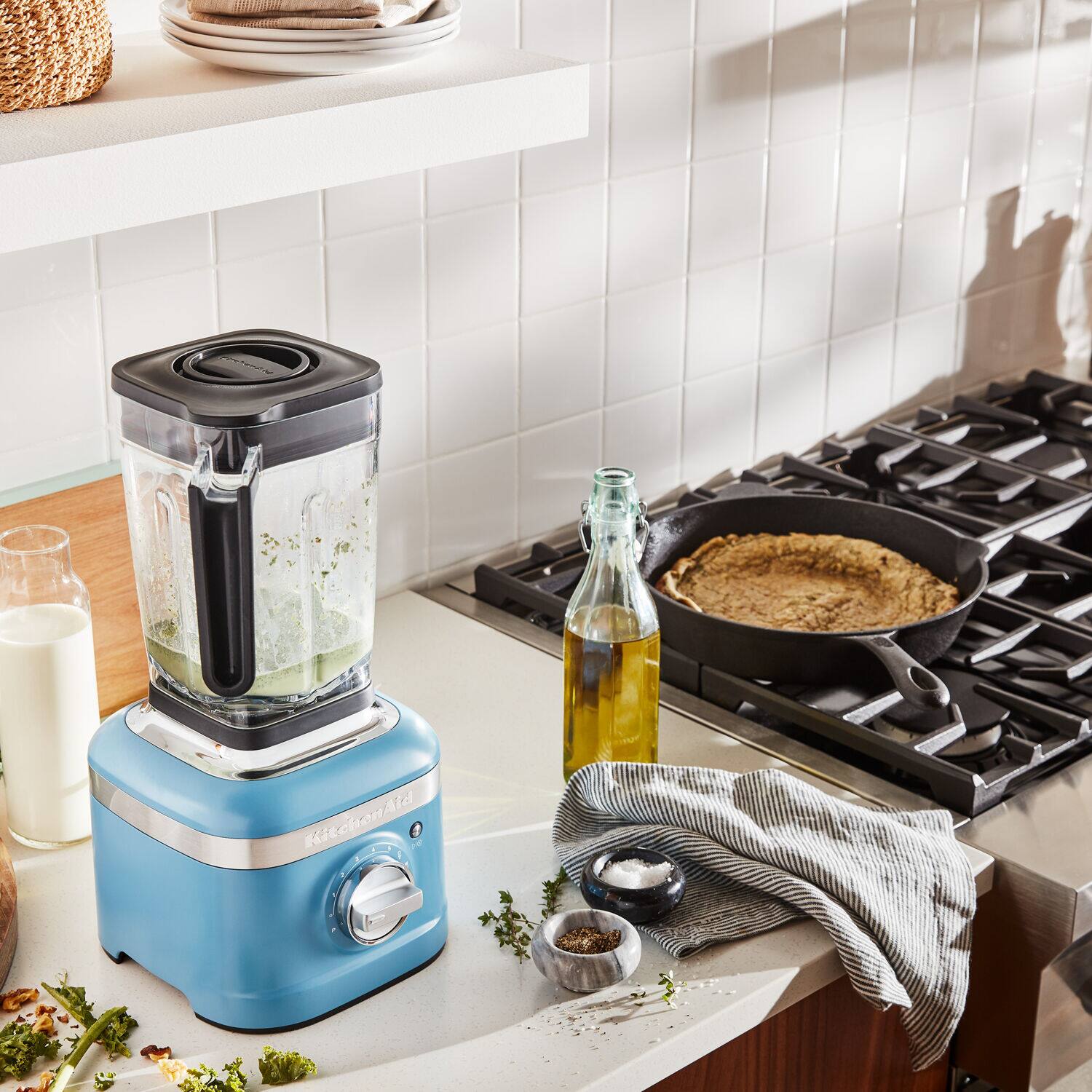 The image shows a kitchen with a blue blender on the counter, a stove, and a bottle of oil. There are also some pots and pans on the stove. The kitchen appears to be well-equipped for cooking and preparing meals.