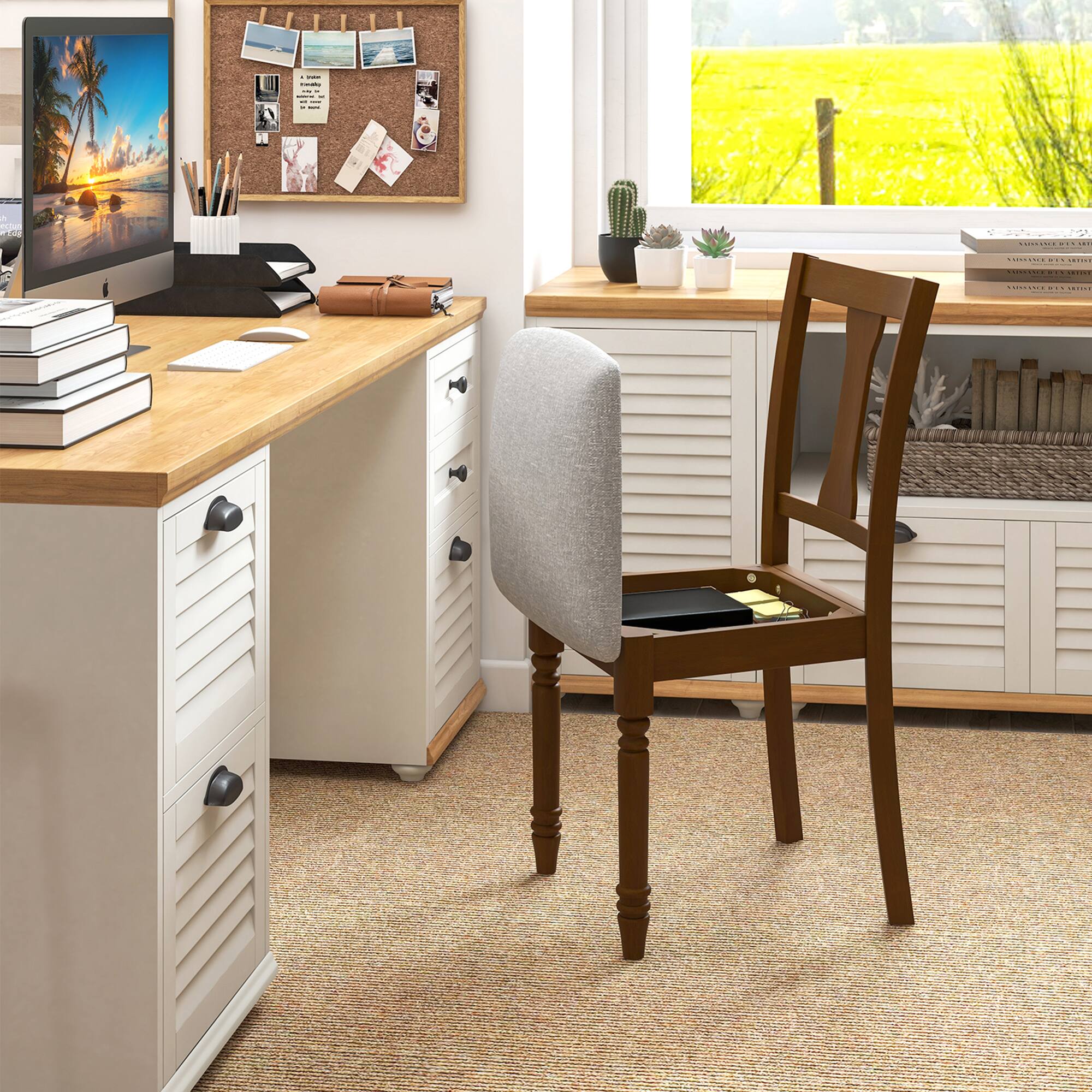 The image shows a well-organized home office setup. A wooden desk with a light-colored finish is positioned against a wall, featuring multiple drawers with black handles. On the desk, there is a computer monitor displaying a beach scene, a stack of books, a pen holder with various stationery items, and a brown leather-bound book. Above the desk, a corkboard is mounted on the wall, adorned with several photos and notes. To the right of the desk, a wooden chair with a gray cushion is placed, and its seat has a small storage compartment. The room has a large window offering a view of a green field, and the floor is carpeted. Additional storage units with similar finishes are visible, holding more books and small potted plants.