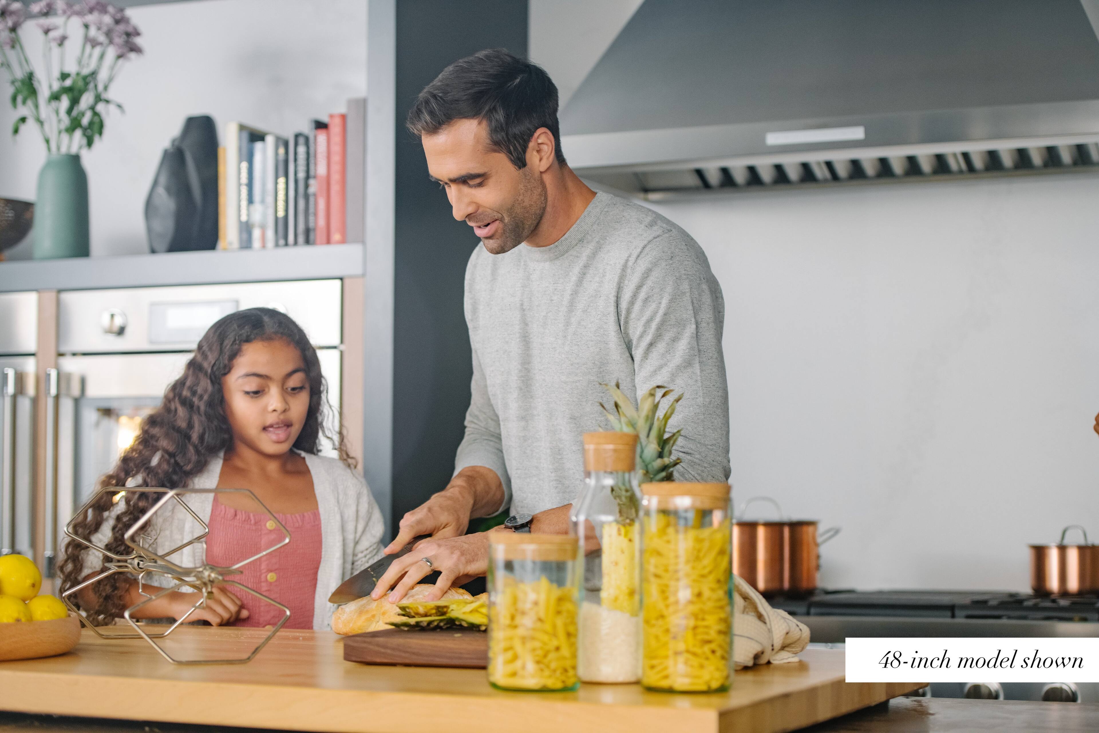 The image shows a man and a young girl in a kitchen, preparing food together. They are standing near a counter with various items on it, including a knife, a bowl, and a couple of bottles. The man is cutting a sandwich, while the girl watches and learns from him. The kitchen is equipped with an oven and a sink, and there are several books visible in the scene, possibly cookbooks or other reference materials. The image is described as a 48-inch model shown, which suggests that it is a representation of a typical kitchen scene, possibly for an advertisement or a design showcase.