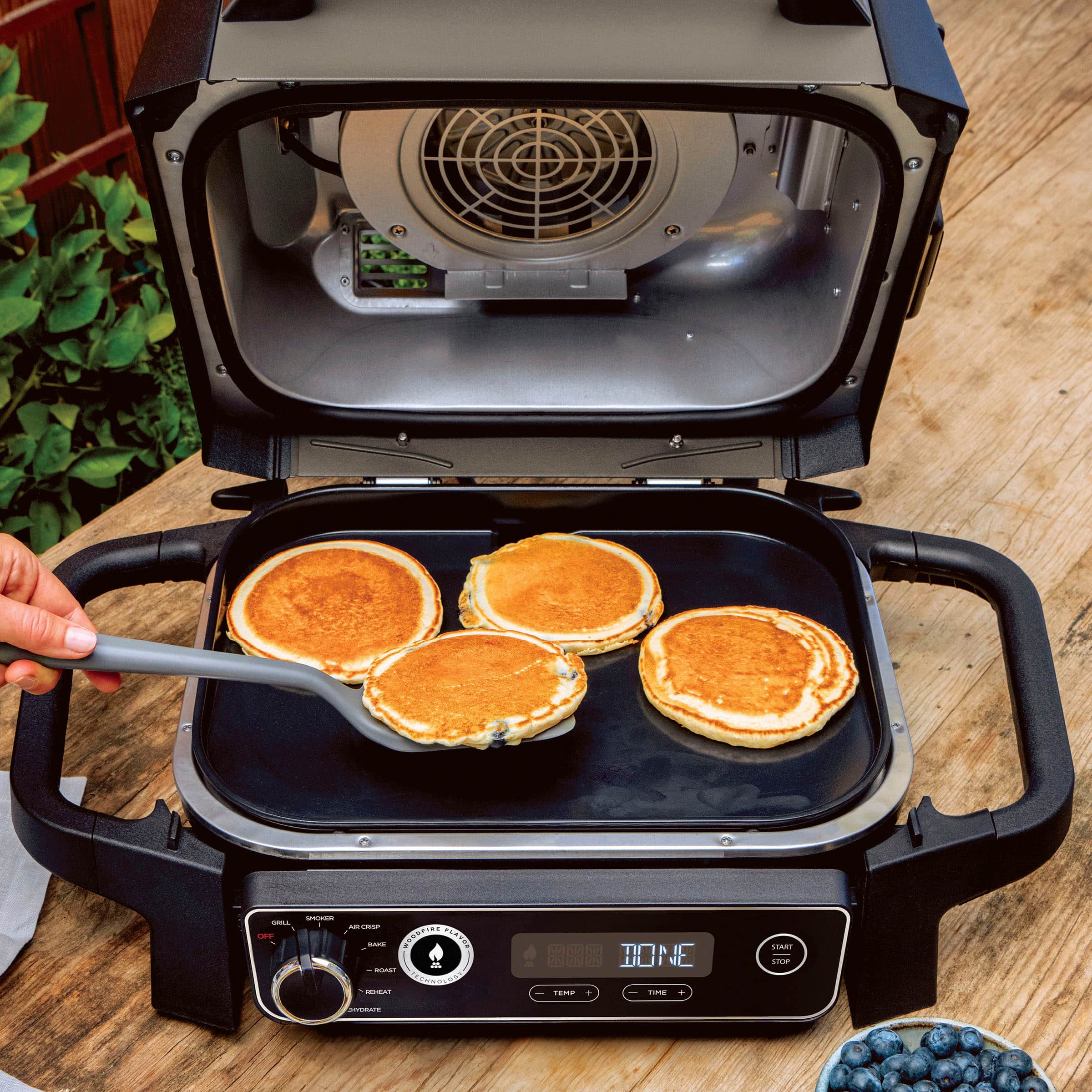 The image shows a person cooking pancakes on a griddle using a toaster oven. The pancakes are being cooked on a stove top, and there are three pancakes visible in the scene. The person is using a spatula to flip the pancakes, ensuring they cook evenly. The toaster oven is placed on the counter, and the pancakes are being cooked to perfection.