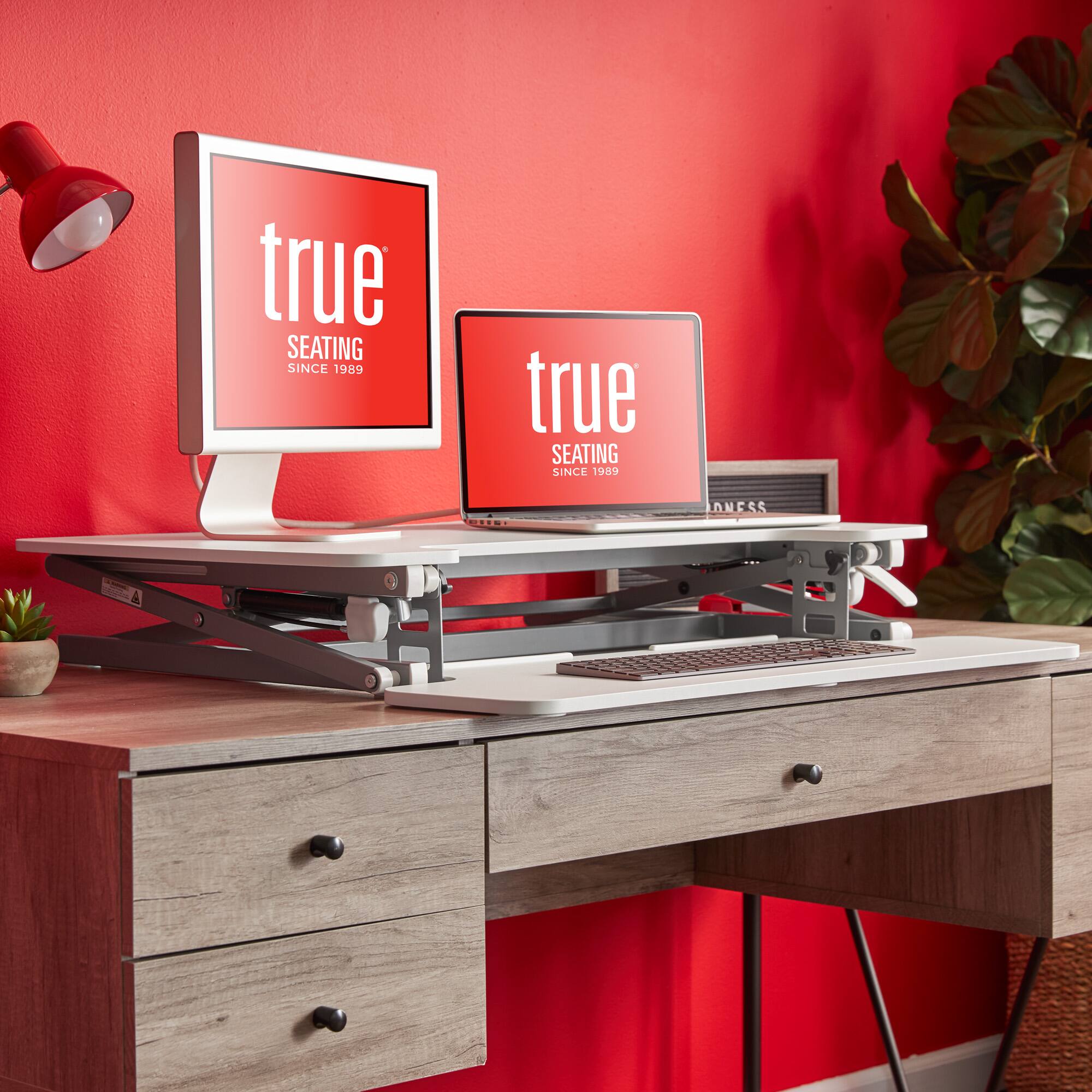The image features a desk with two laptops, one on the left and one on the right, placed on a stand. The desk is situated in front of a red wall, and there is a potted plant nearby. The laptops are placed on a white desk, which is a part of a computer workstation. The desk is also equipped with a keyboard and a mouse, making it a functional workspace.