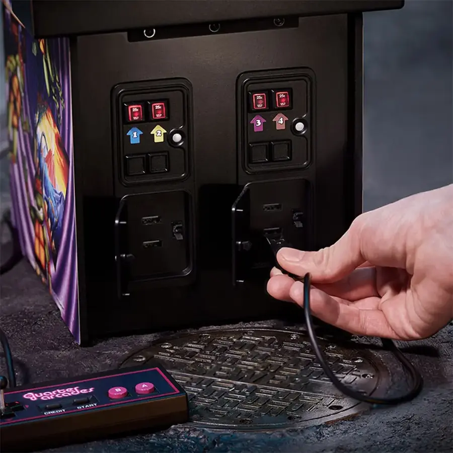 The image shows a person holding a black wire and plugging it into a black box with a bunch of switches on it. The box is located on a table. The person is using the black wire to connect the box to a game console. The game console is placed on the table next to the box.