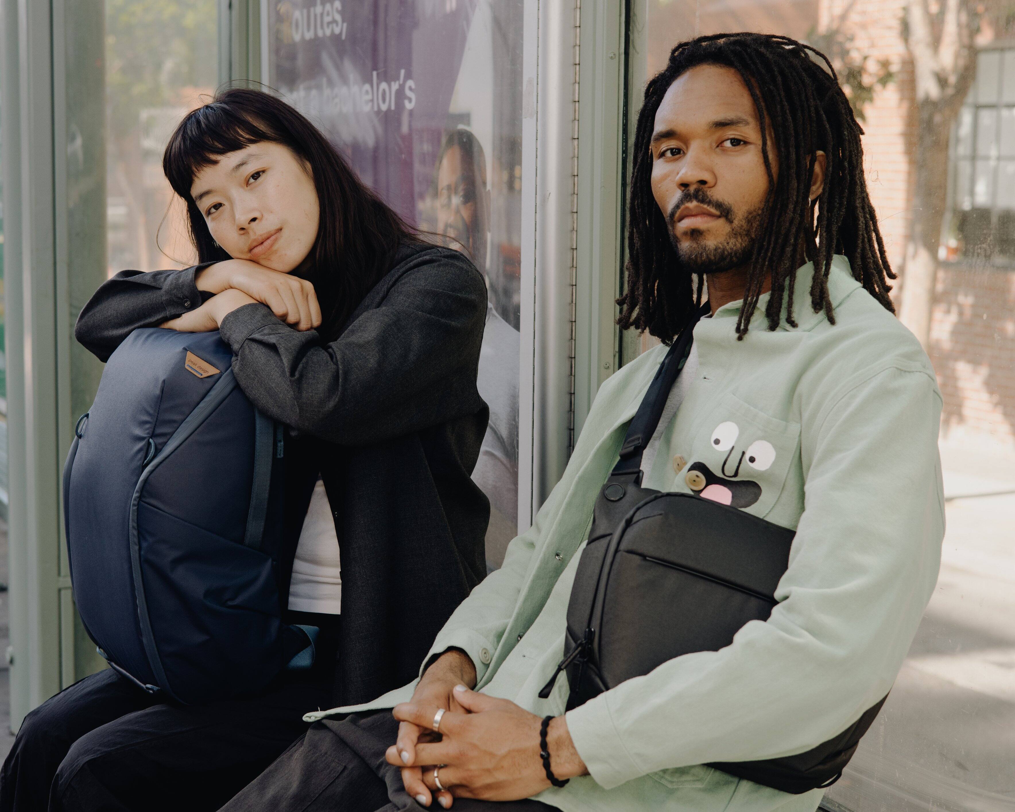 Outdoors, a young man and a young woman are sitting on a bench. The man is wearing a backpack and a strap across his chest, while the woman has a backpack on her lap. They seem to be waiting for something or someone, possibly a bus or a friend. The image captures a moment of rest and companionship between the two individuals.