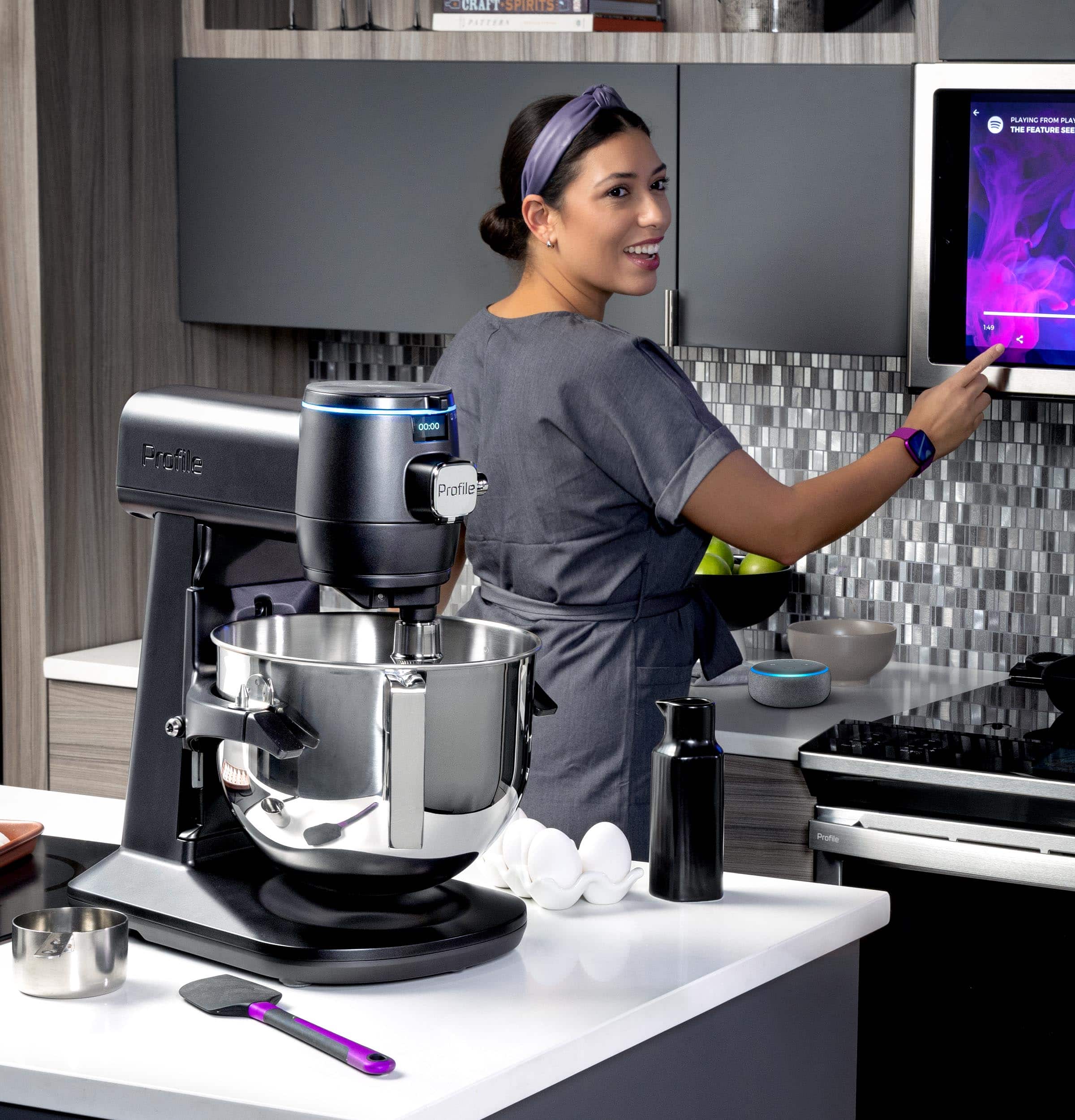 The image features a woman standing in a kitchen, preparing food. She is using a mixer to blend ingredients, and there are multiple bowls on the countertop. The woman is smiling, indicating that she is enjoying the process. The kitchen is well-equipped with various appliances, including a microwave and an oven. There are also several books and bottles scattered around the kitchen, possibly containing recipes or ingredients.