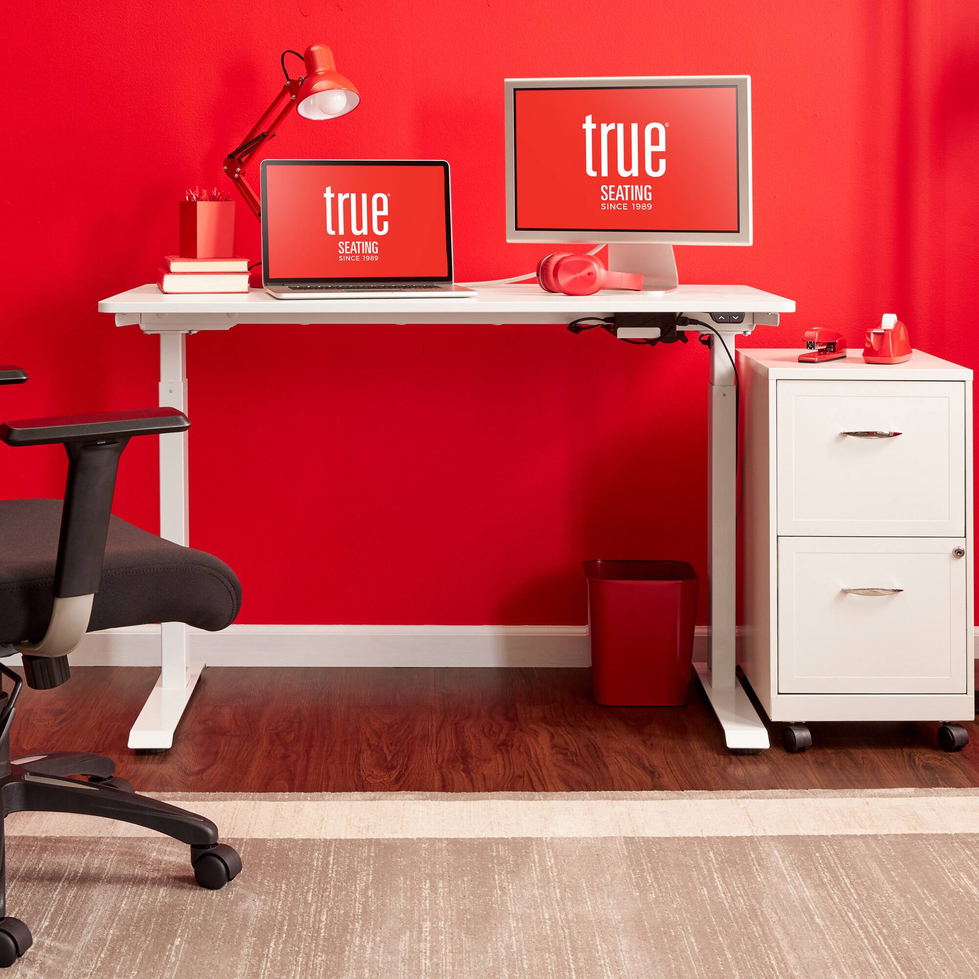 The image features a red desk with a laptop, a monitor, and a keyboard. There is also a red chair positioned in front of the desk. The desk is adorned with a red wall, giving the space a bold and vibrant appearance. The desk is equipped with a lamp, which provides lighting for the workspace. Additionally, there is a book placed on the desk, suggesting that the user may be engaged in reading or studying.