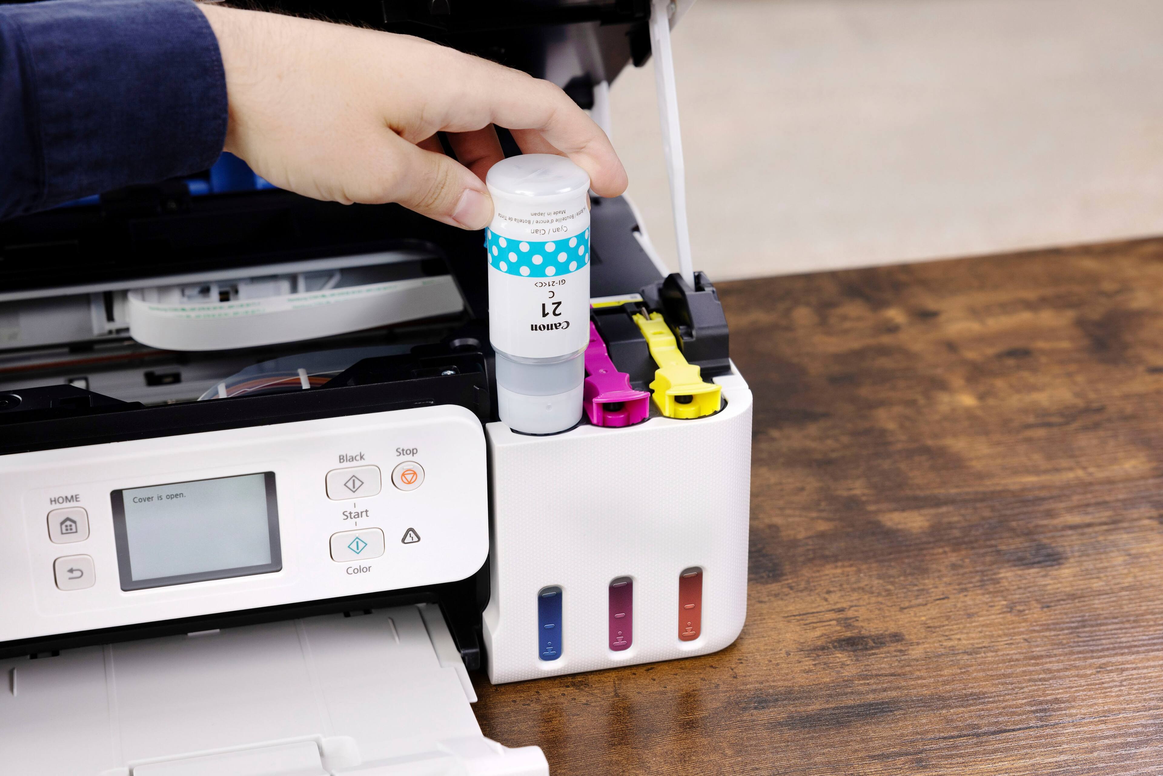The text on the image reads: "AN - - A... A - mURTE nR R Ciar / Cyan 1 21 canon Black Stop HOME Cover i open I - Start - I Color." This text is likely a description or instructions for using the printer, with the person holding the bottle of ink to fill the printer.