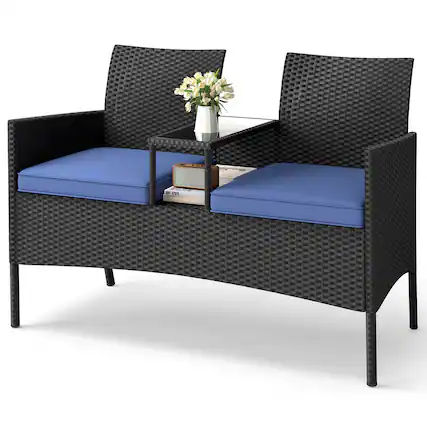 The image shows a black wicker chair with a blue cushion on it. The chair is placed next to another chair, and there is a table in between them. On the table, there is a vase with flowers in it, adding a touch of elegance to the scene. Additionally, there is a book on the table, suggesting that someone might be reading or studying in this cozy setting.