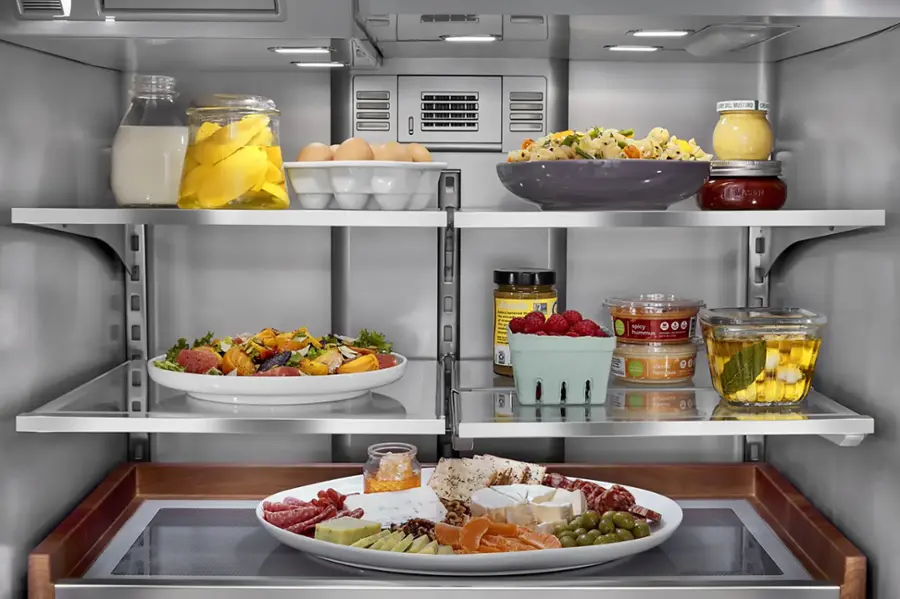 The refrigerator is stocked with a variety of food items, including a bowl of fruit, a bowl of vegetables, and several jars of condiments. There are also multiple bottles of condiments, such as mustard and hummus, as well as a container of strawberries. The refrigerator is well-organized, with the food items placed on shelves and in drawers. The presence of these items suggests that the refrigerator is well-stocked and ready for use.