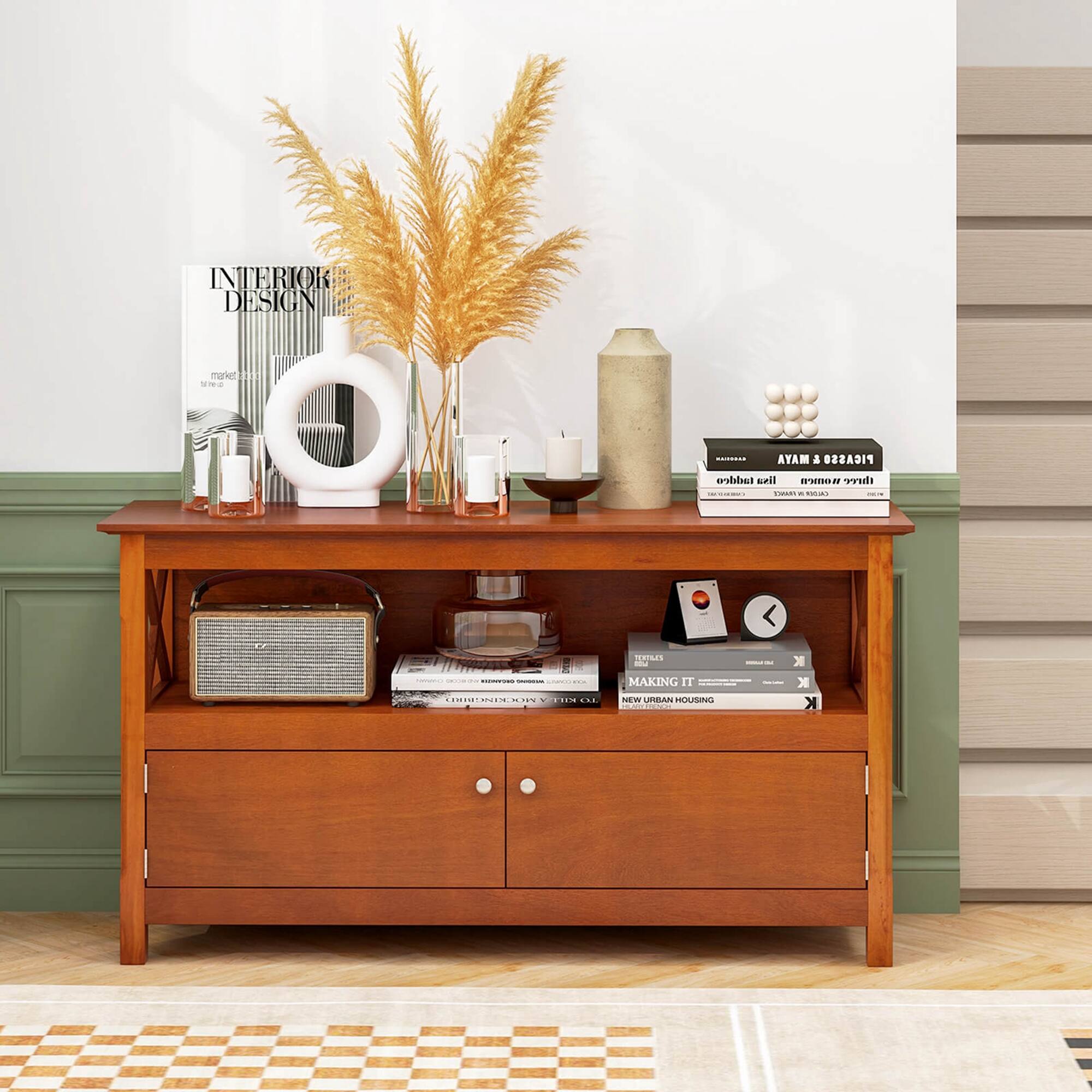 The image features a wooden table with various items on it, including a vase of wheat, a candle, and books. The table is placed in front of a wall, and there is a bookshelf nearby. The table is also decorated with a clock and a bowl. The scene appears to be a cozy and well-organized living space, with the table serving as a focal point for the room.