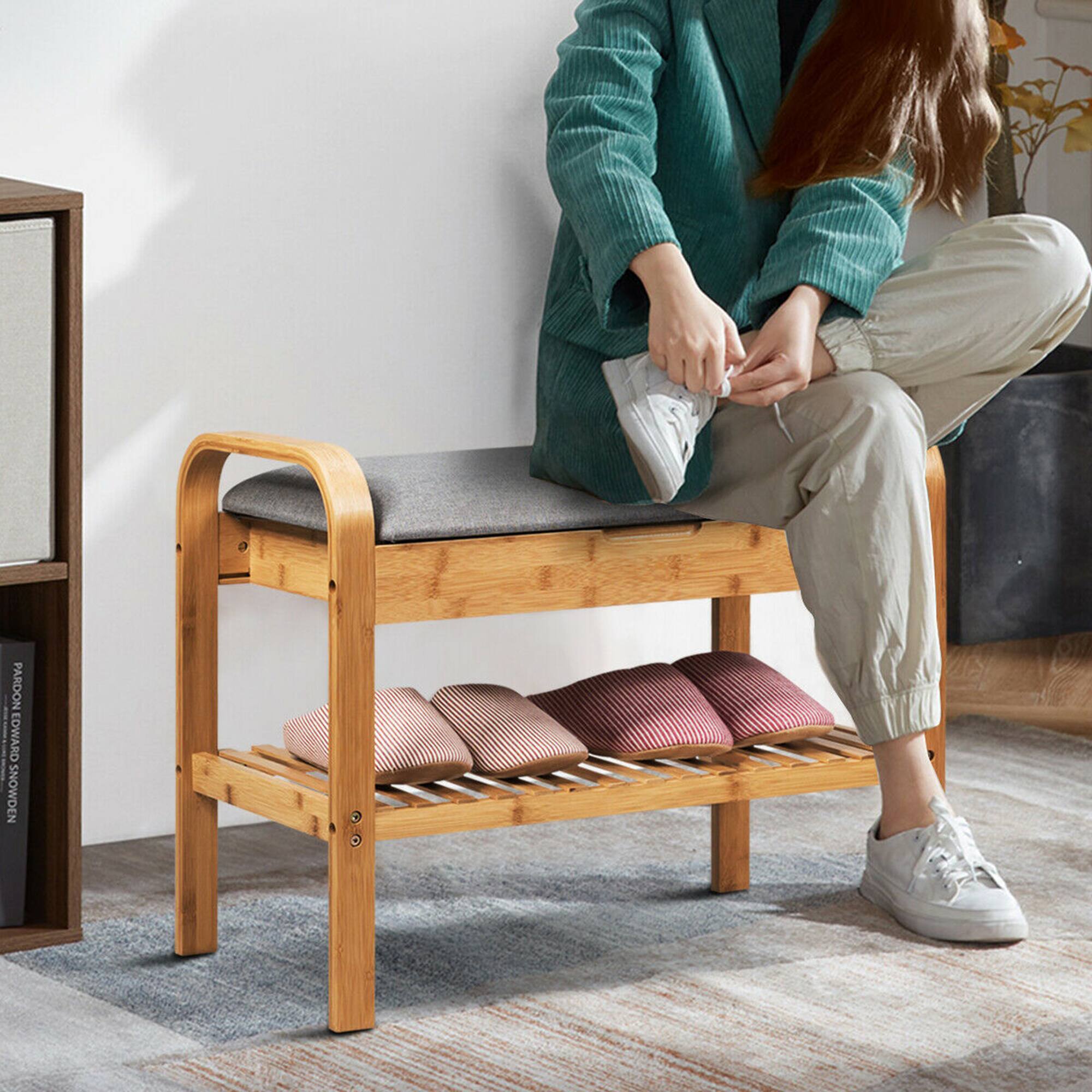 The image shows a woman sitting on a wooden bench in a room. She is wearing a green jacket and appears to be tying her shoes. The bench has a shelf underneath it, and there are several books on the shelf. Additionally, there is a potted plant in the room, and a vase is placed nearby. The woman seems to be focused on her task, and the overall atmosphere of the scene is calm and relaxed.