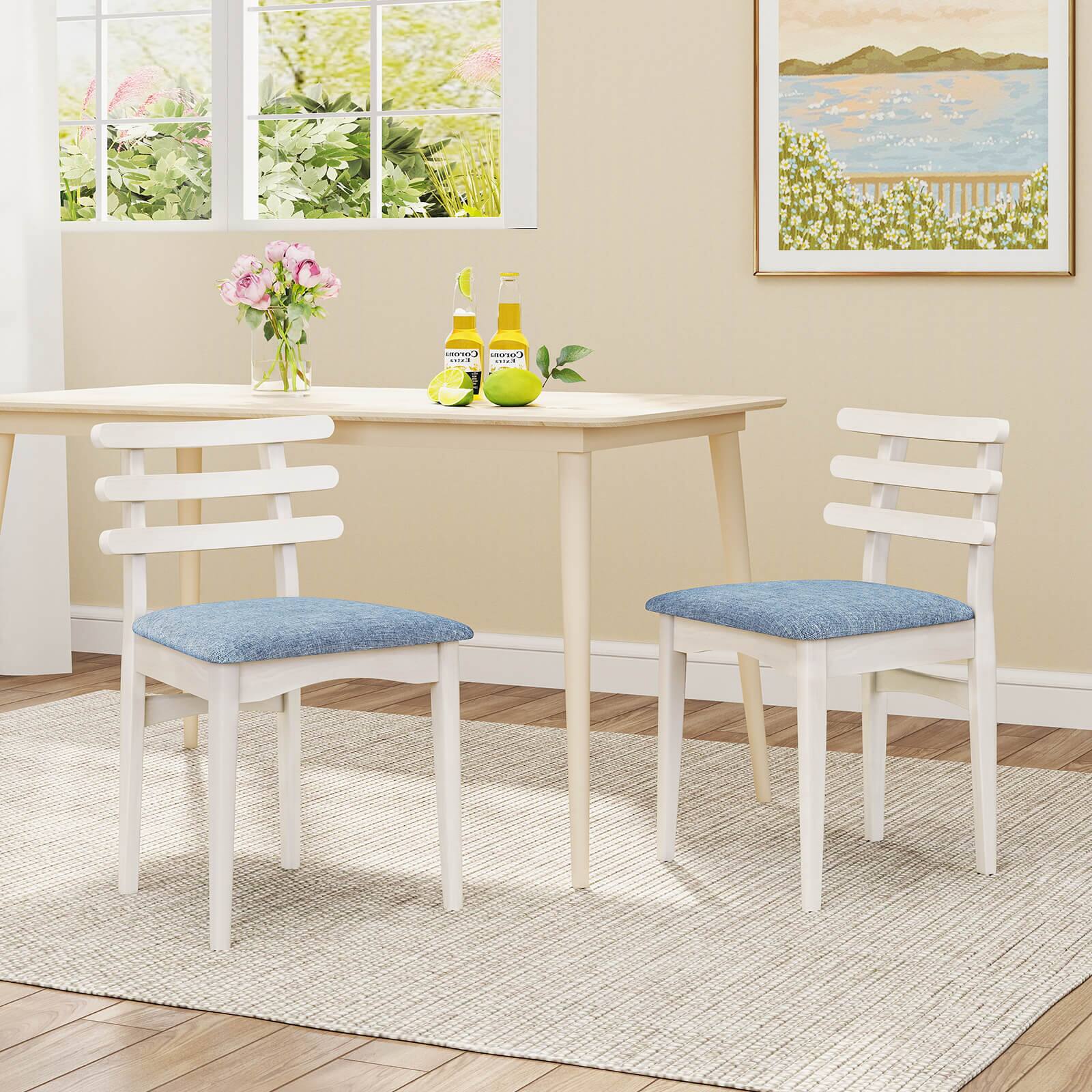 The image shows a dining table with four chairs around it. The table is set with two bottles of beer and a vase of flowers. The chairs are white and blue, and the table is white. The table is positioned near a window, providing natural light to the room. The overall scene creates a pleasant and inviting atmosphere for a meal or gathering.